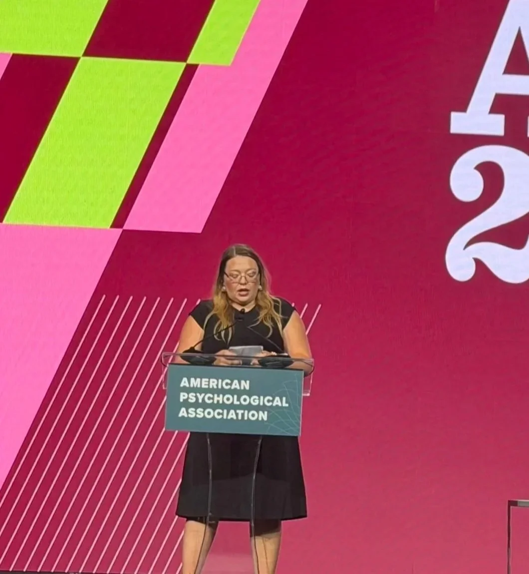 A woman speaking at a podium with a sign reading 'American Psychological Association' at a conference or event, with a colorful background featuring geometric designs.