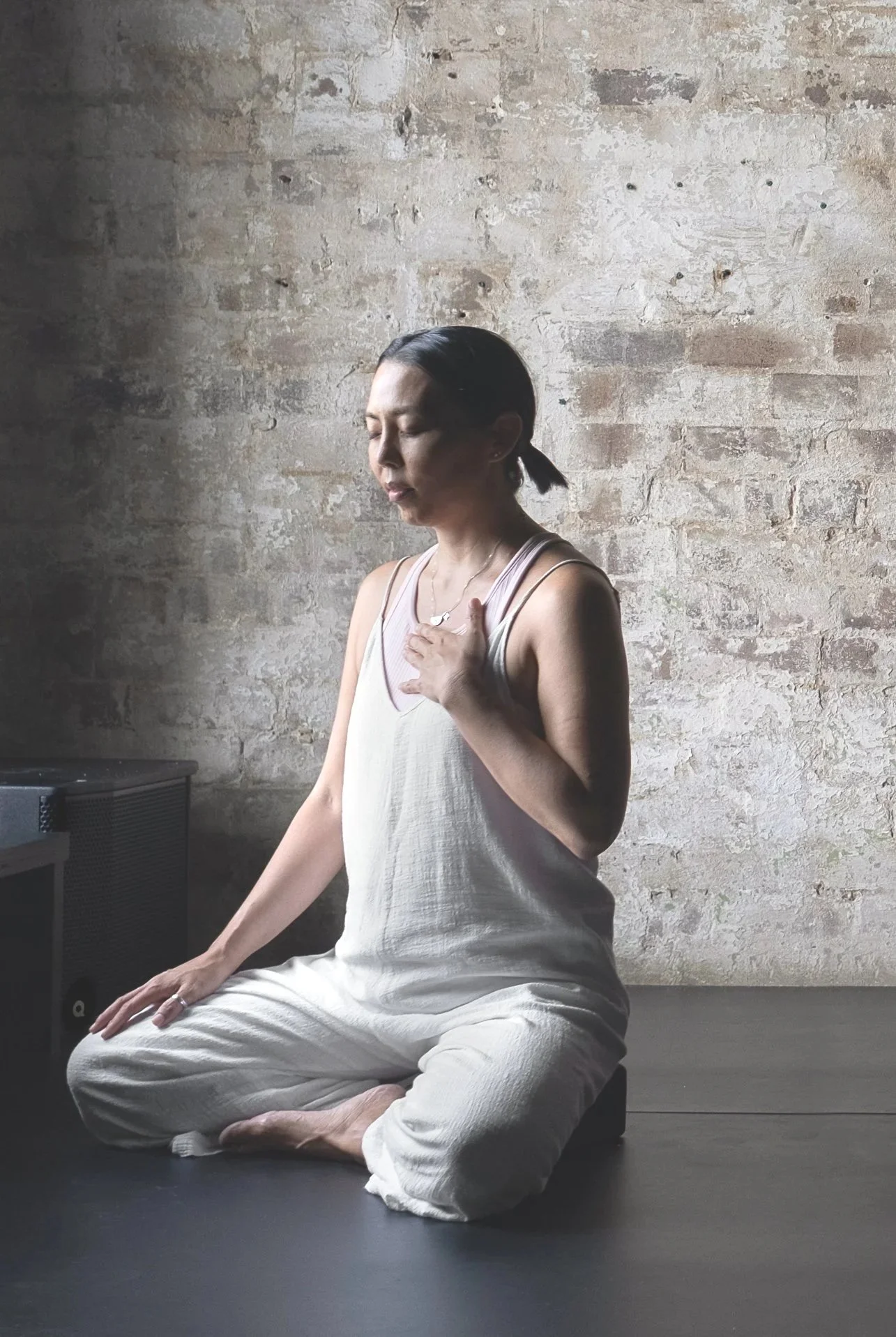 Woman practicing meditation or yoga in a cross-legged pose against a brick wall with her hand on her chest and eyes closed.