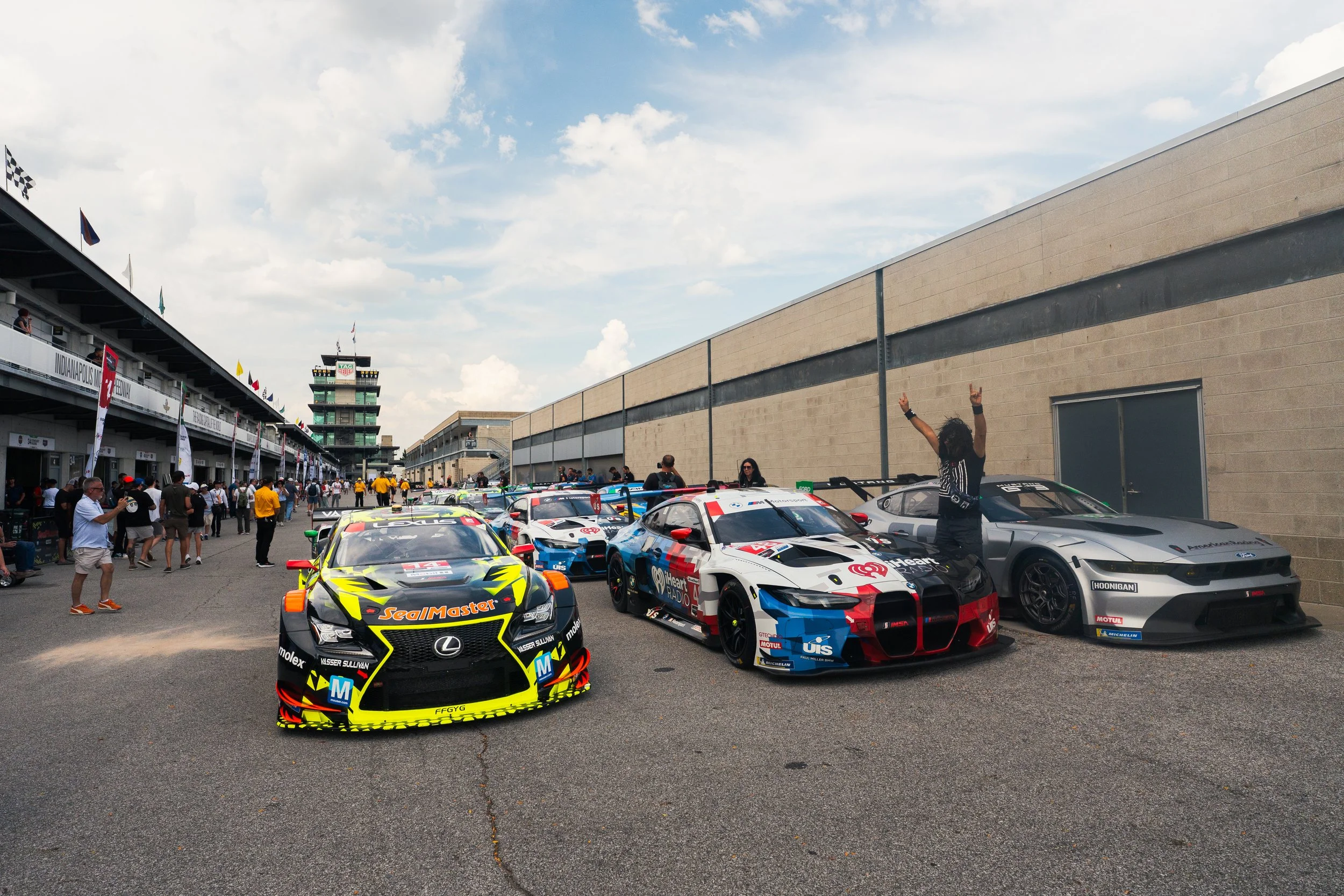 Race cars parked near a pit wall at a racetrack with people walking around and a tower in the background.