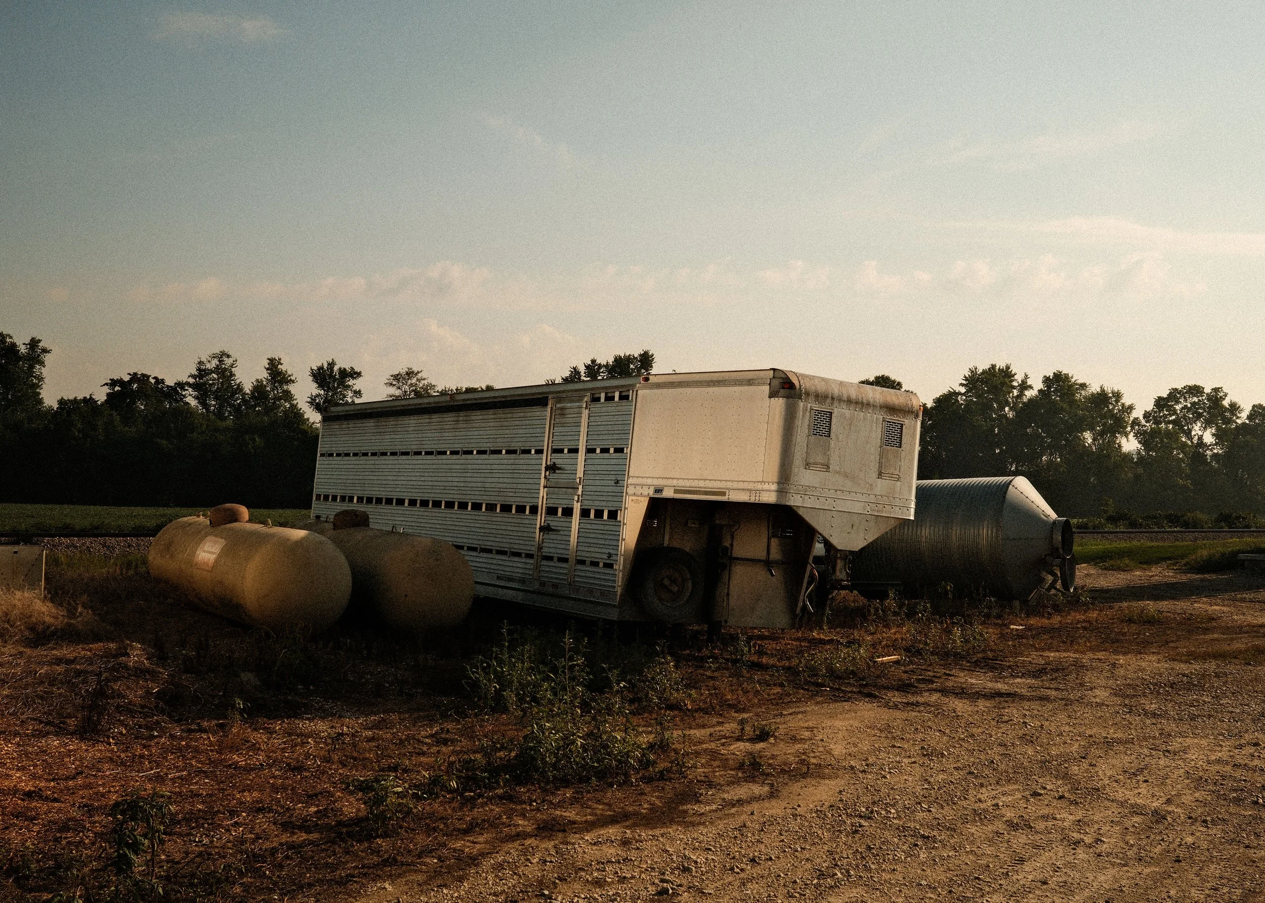 An abandoned livestock trailer and large tanks on a dirt field at sunset with trees in the background.