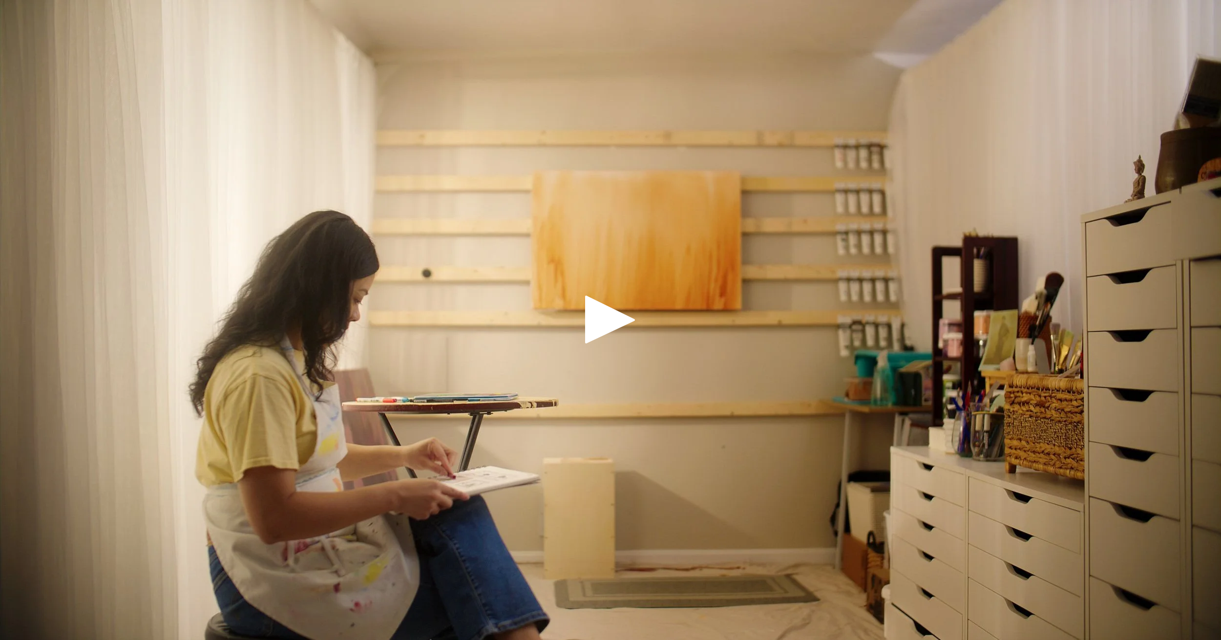 A woman sitting in a room with beige curtains, looking down at a book or notebook. The room has shelves with supplies, drawers, and wall-mounted hooks or small shelves in the background.