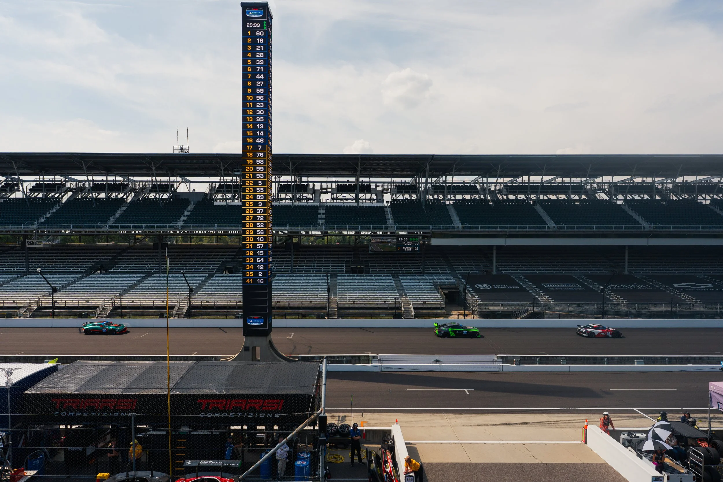 A race track at a motorsport event with three race cars on the track, a tall scoreboard displaying qualifying or race times, and empty grandstands in the background under a partly cloudy sky.