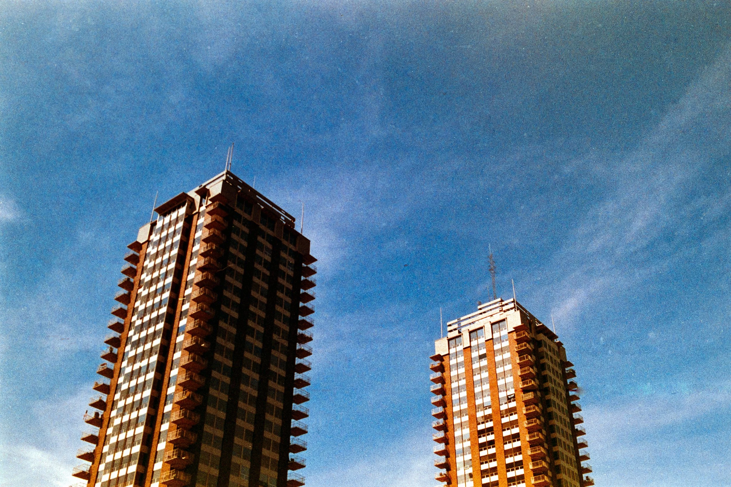 Two tall modern buildings with multiple balconies against a blue sky with wispy clouds.