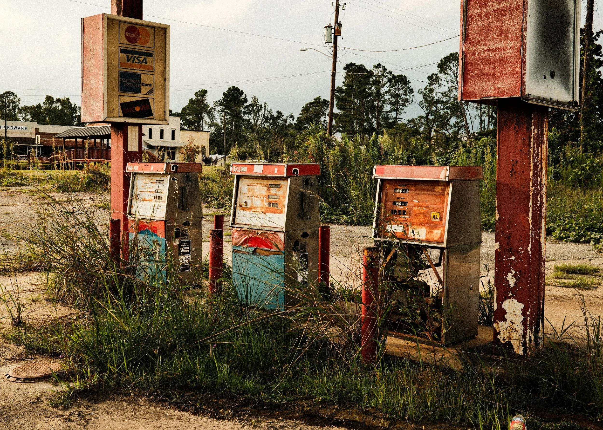 Three old, rusty, and damaged payphone booths beside a weathered red sign and a utility pole, with overgrown grass and a cloudy sky in the background.