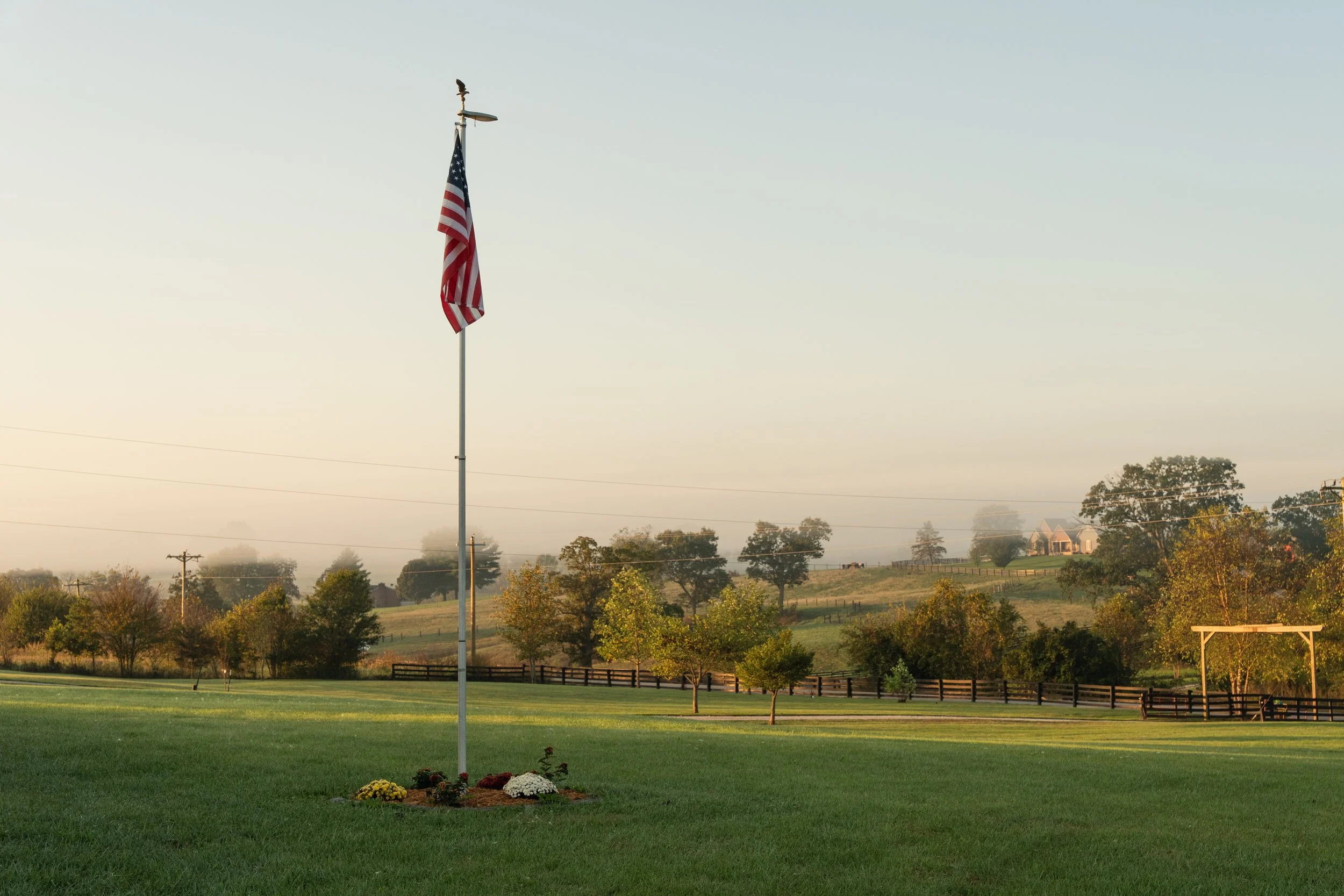 American flag flying on a flagpole in a grassy field with a small flower bed in the foreground, trees in the background, and a misty hillside with houses.