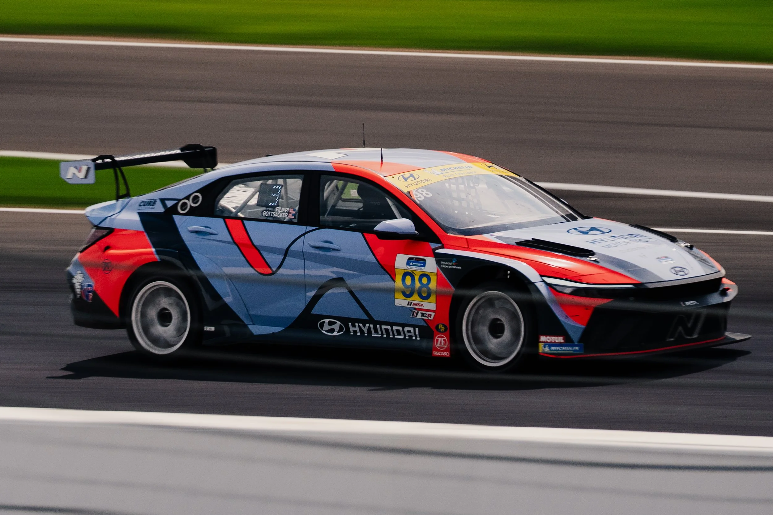 A race car on a track with a colorful livery featuring Hyundai and Michelin logos, number 98, and a large rear wing, moving at high speed.