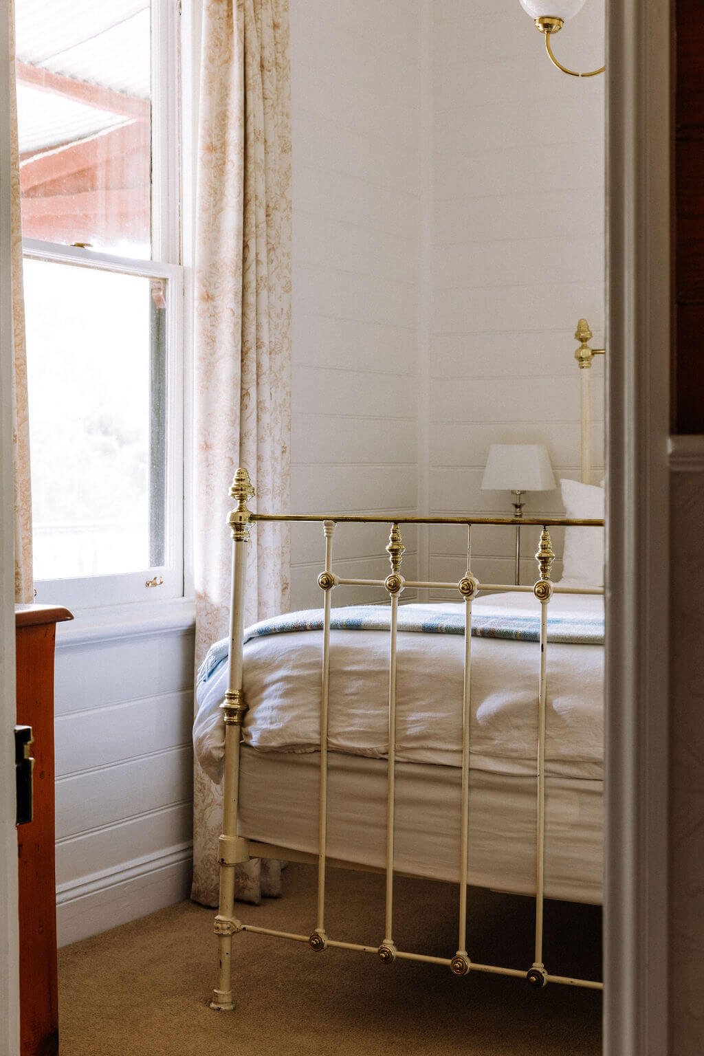 The Inn Beechworth, Luxury Accommodation - Kelly House. Cozy bedroom with a vintage brass bed frame, white linens, and a bedside lamp next to a window with patterned curtains.