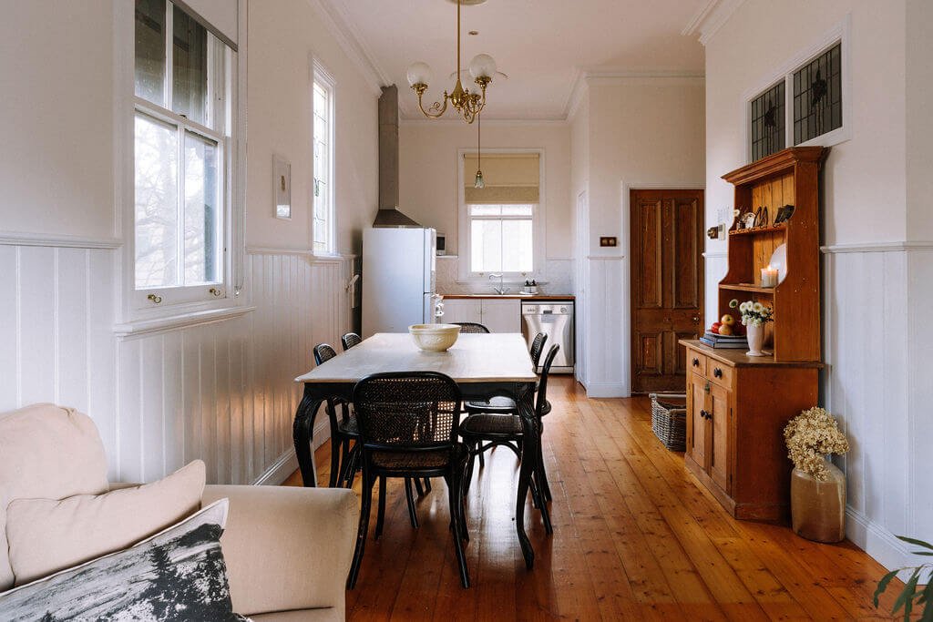 The Inn Beechworth, Luxury Accommodation. Cozy kitchen and dining area with wooden floors, a table with chairs, a hutch with decor, and a view of the kitchen appliances like a fridge and stove.