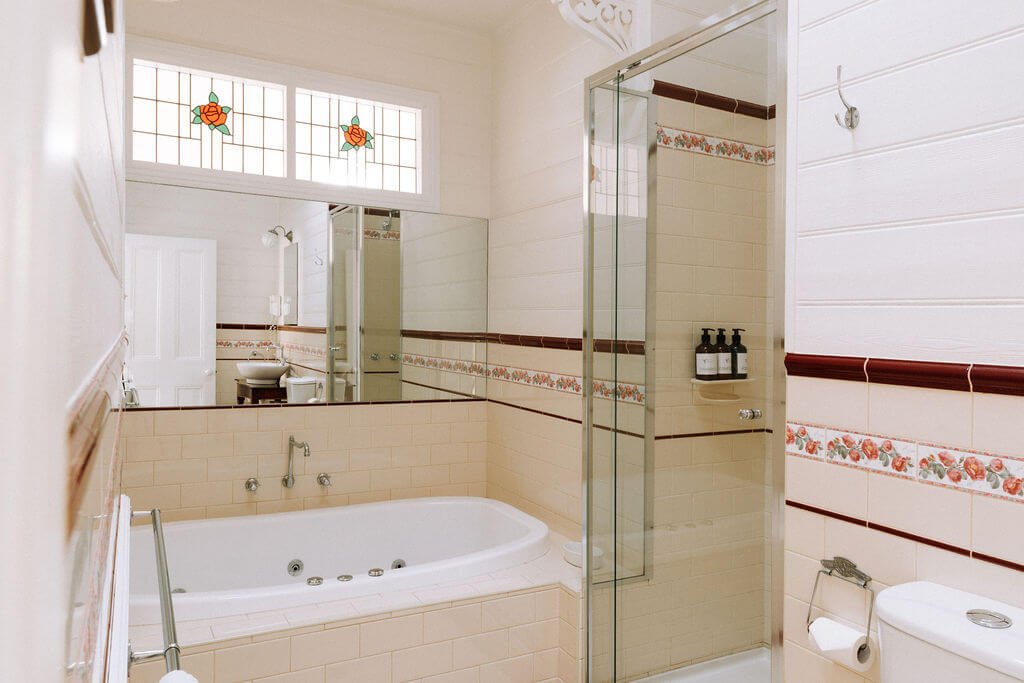 The Inn Beechworth, Luxury Accommodation - Kelly House. Vintage-style bathroom with beige tiles, floral accents, a large bathtub, glass shower door, stained glass window, and toiletries on a shelf.