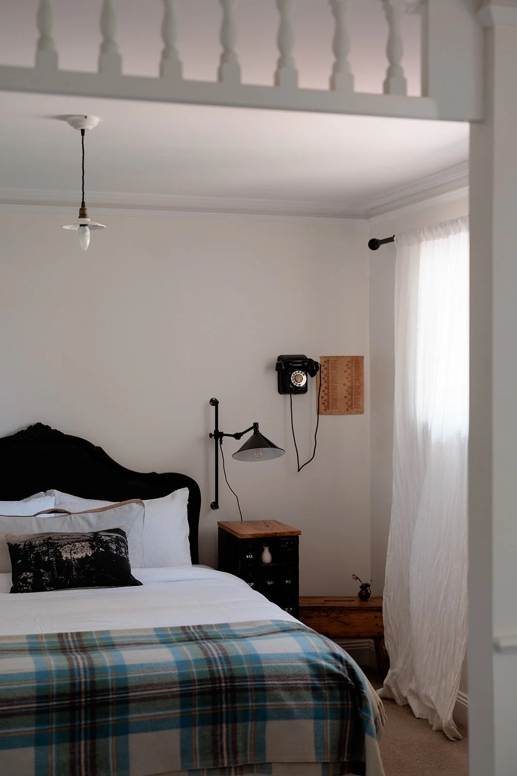 Picketts Cottage has a cosy bedroom with a black headboard, white bedding, and a plaid blanket. There's a wooden nightstand, a wall-mounted swing-arm lamp, a vintage black rotary phone, and white curtains letting in natural light.