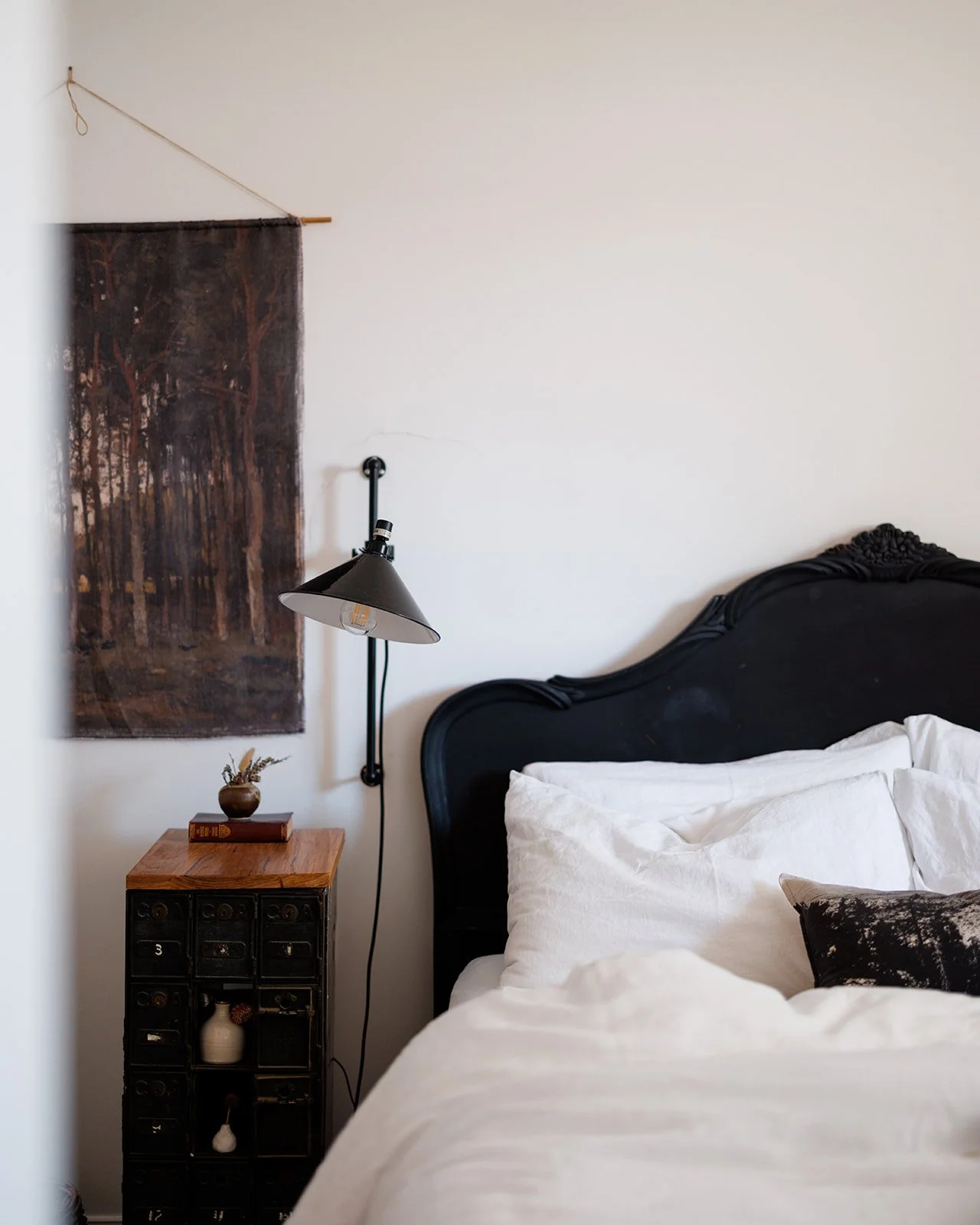 Pickett's Cottage has a cosy bedroom with a black ornate headboard, white bedding, a wall-mounted reading lamp, a small wooden side table with decorative items, and a dark forest landscape tapestry hanging on the white wall.