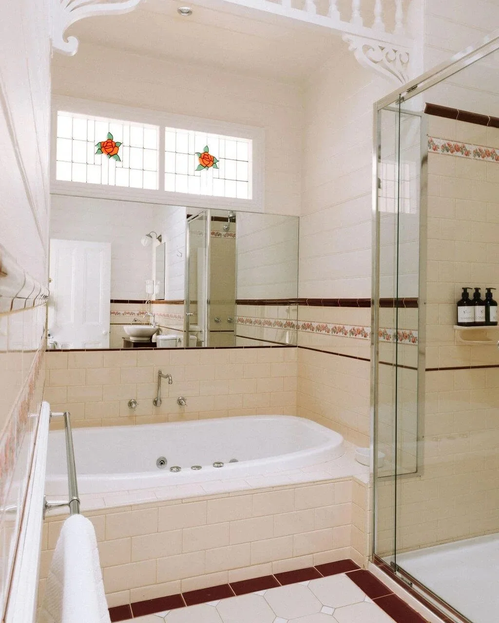Bathroom with a bathtub, a glass shower enclosure, a wall mirror, a window with stained glass roses, and beige tiles with floral border accents.