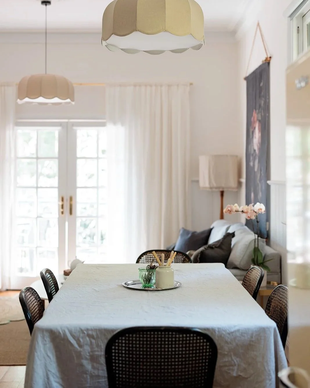 Dining room with a table covered by a light-colored cloth, surrounded by black rattan chairs. The table has a tray with a white jar and a green glass. In the background, there is a sofa with gray pillows, a vase with pink flowers, a framed artwork, a