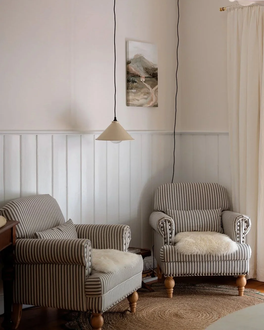 Living room corner with two striped armchairs, a wall-mounted painting of mountains, a hanging lampshade, cream-colored curtains, a small side table, and a round rug.