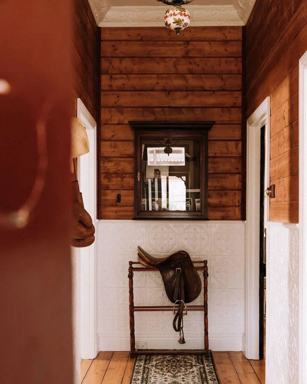 The Inn Beechworth. Luxury Accommodation - Kelly House Hallway with wooden walls, saddle on a rack, patterned rug, and hanging light fixture.