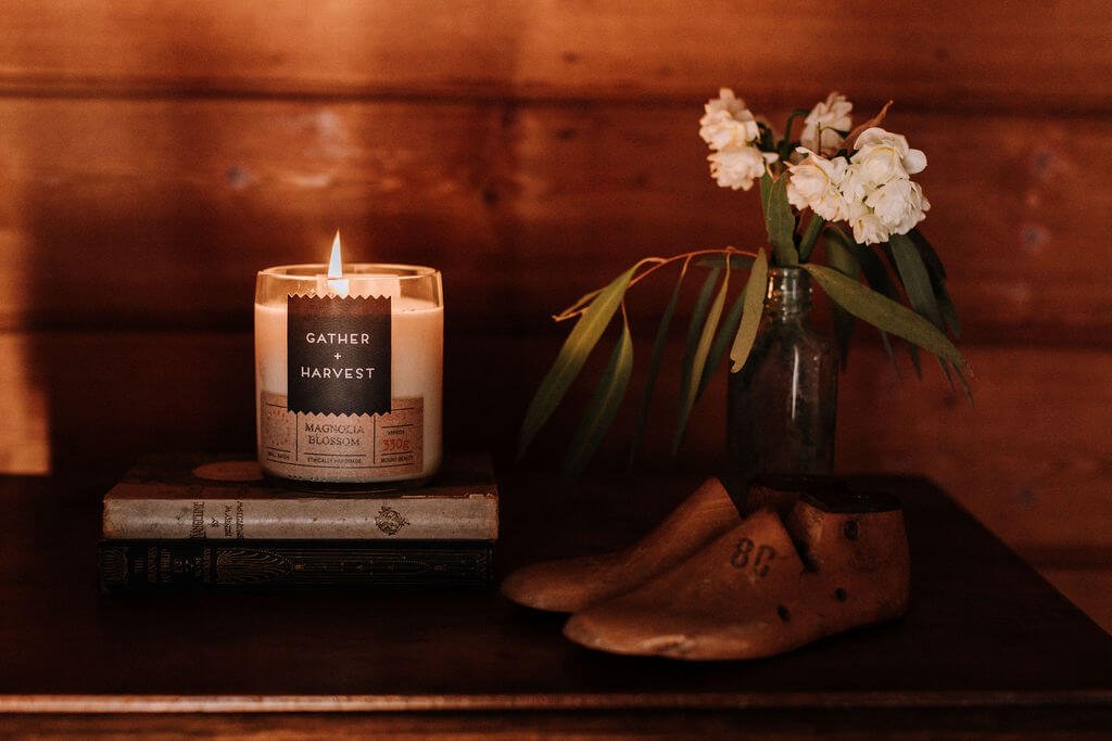 The Inn Beechworth, Luxury Accommodation - Kelly House. Lit candle labeled "Gather + Harvest" on stacked books, vintage shoe lasts, flowers in glass vase on wooden surface.