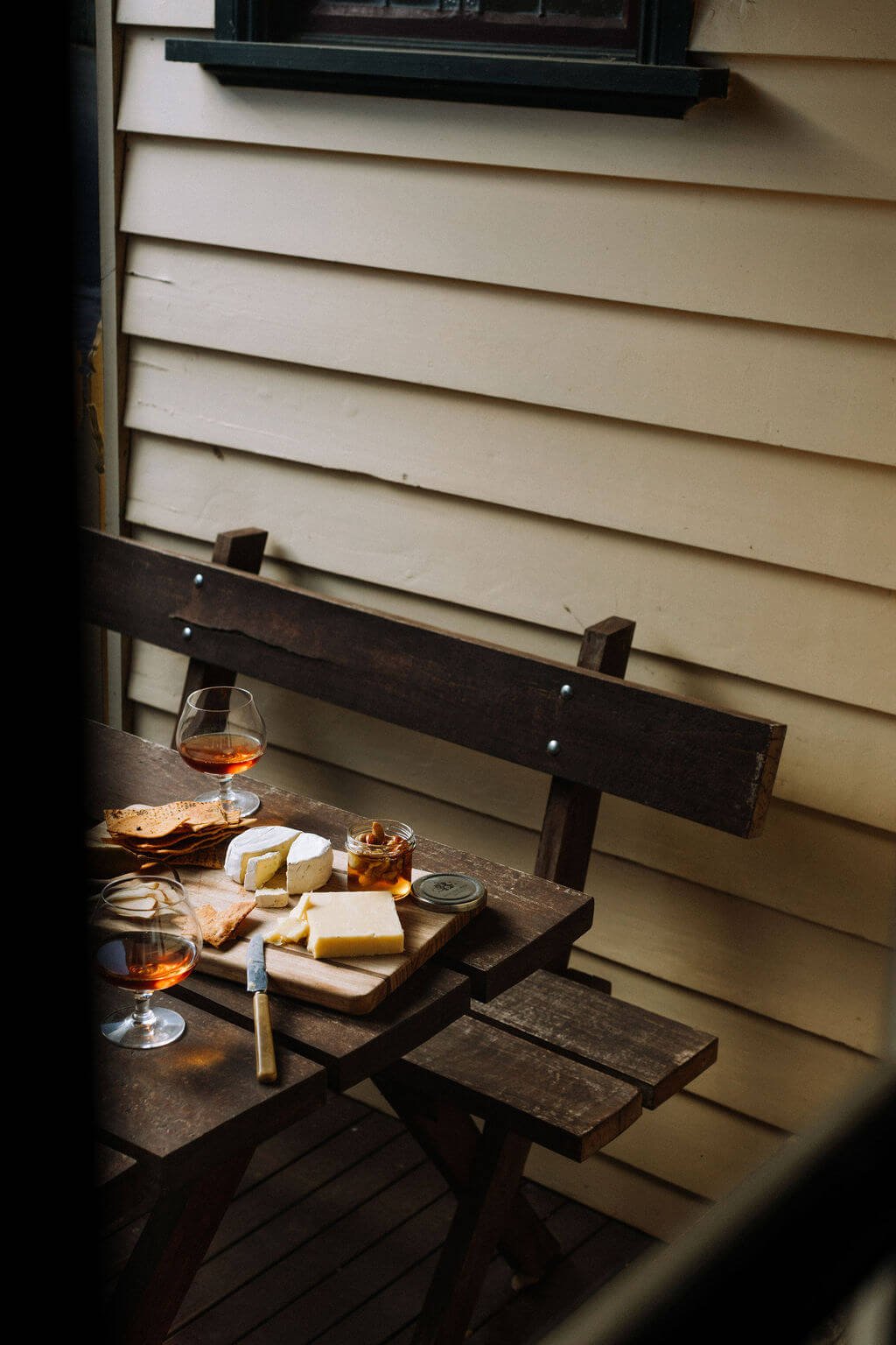 The Inn Beechworth, Luxury Accommodation - Kelly House. Rustic wooden table with cheese platter, crackers, and two glasses of rosé wine on a patio bench.