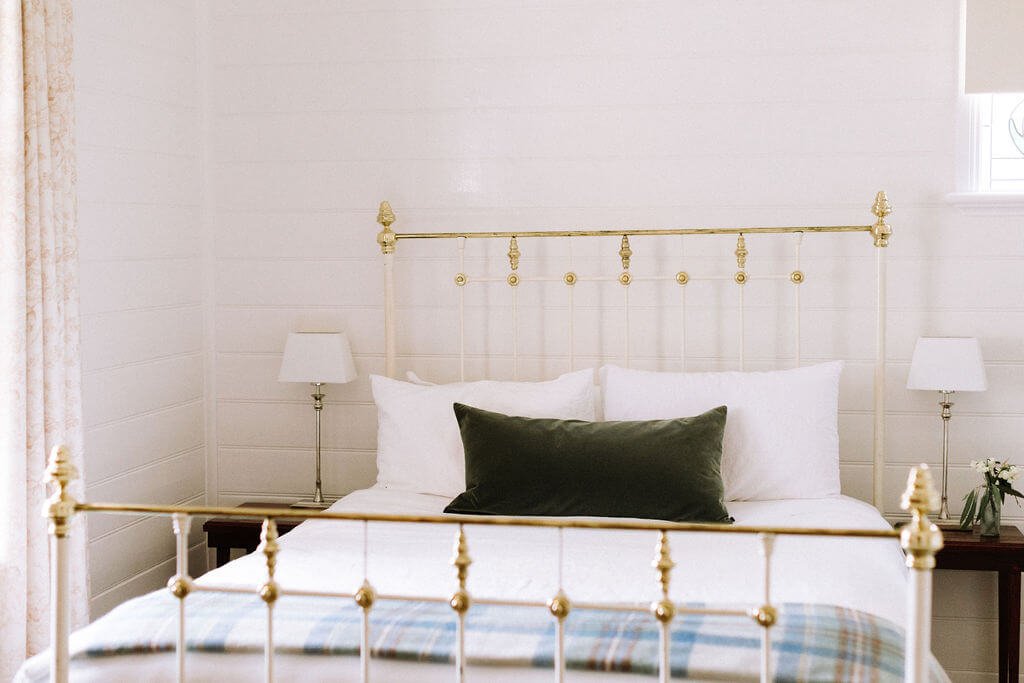 The Inn Beechworth, Luxury Accommodation - Kelly House. Cozy bedroom with brass bed frame and decorative pillows, white walls, and bedside tables with lamps.