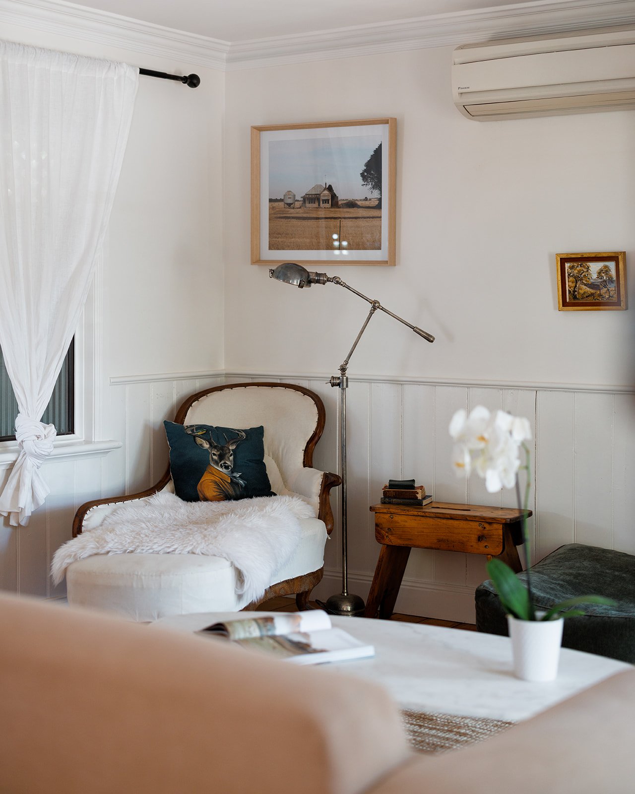 Pickett's Cottage living room corner with a white armchair, a throw pillow, a small wooden side table, framed artwork on the wall, a floor lamp, a potted white orchid, and a window with white curtains.