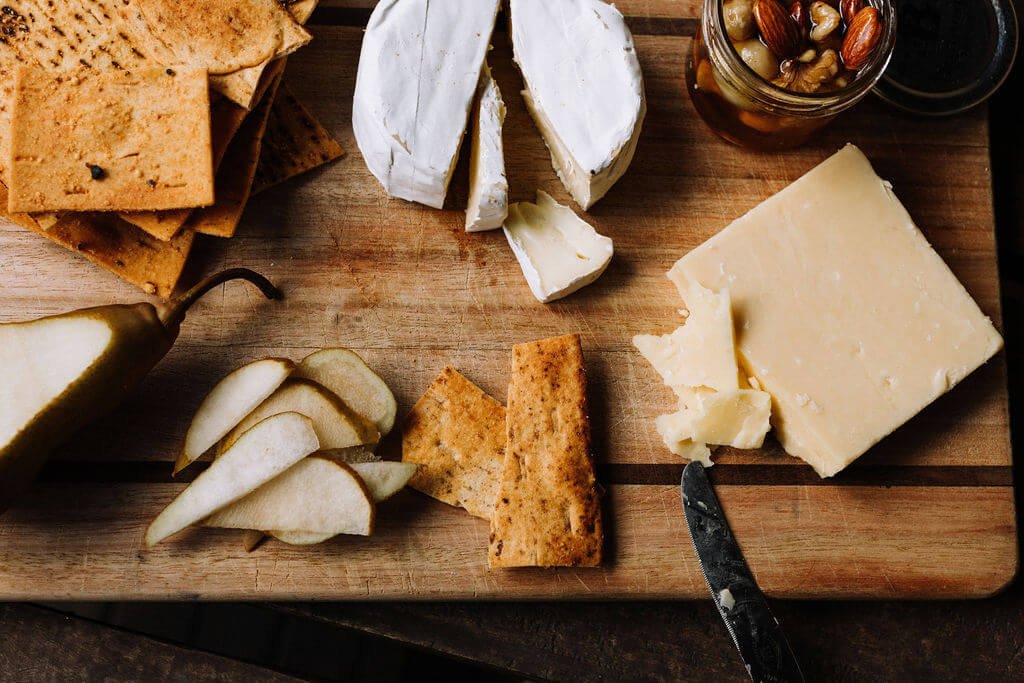 The Inn Beechworth, Luxury Accommodation - Kelly House. Cheese board with assorted cheeses, sliced pear, crackers, honey jar with nuts, and a knife on a wooden surface.