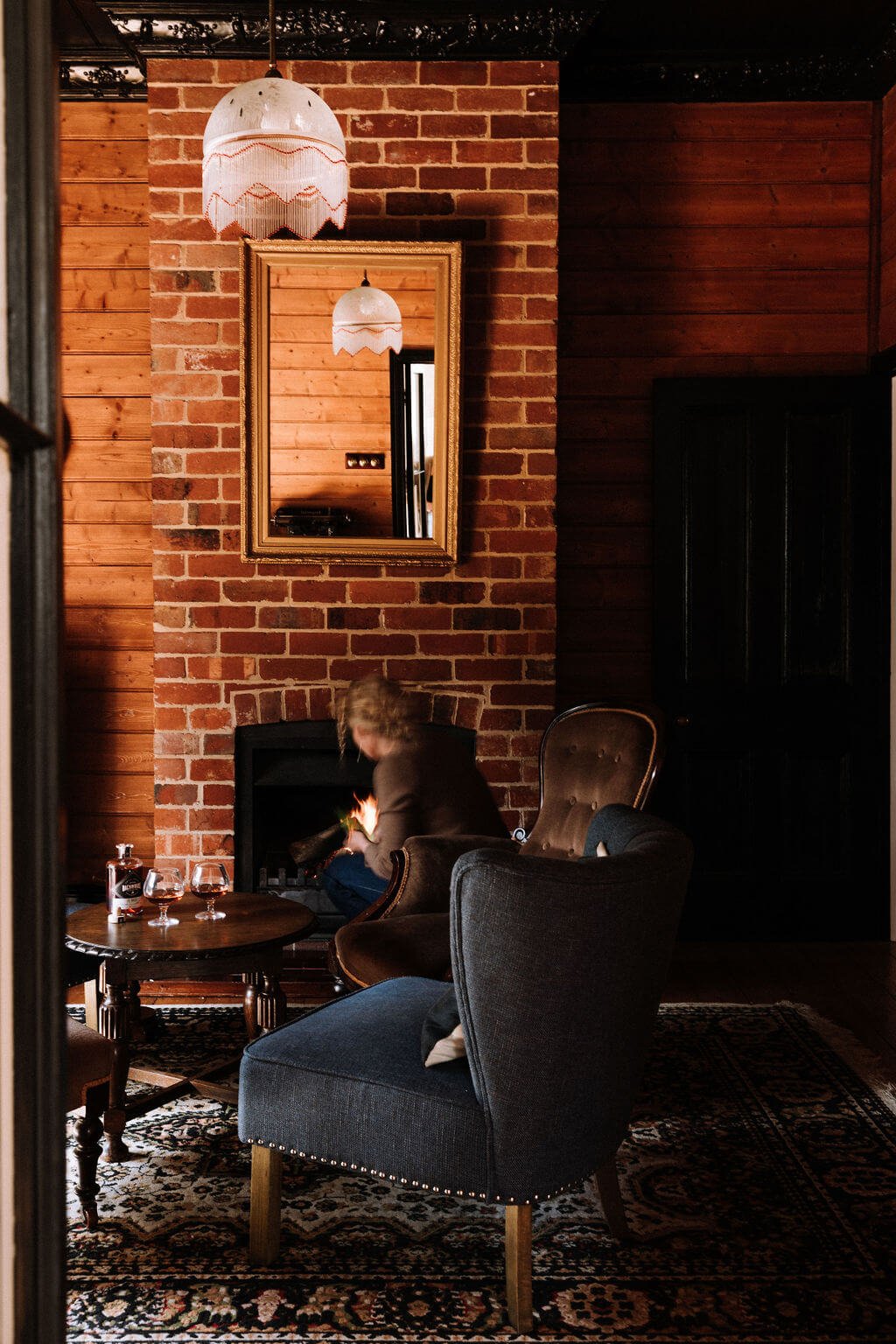 The Inn Beechworth, Luxury Accommodation - Kelly House. A cozy living room with a person tending a fireplace, chairs, a wooden table with glasses and a bottle, an ornate rug, and a mirror reflecting a hanging lamp.