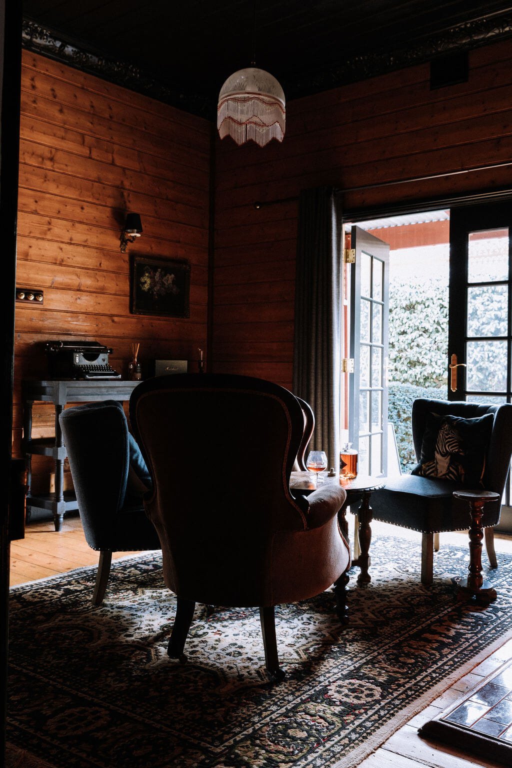 The Inn Beechworth, Luxury Accommodation - Kelly House. Cozy wood-paneled room with vintage furniture, including a typewriter on a table, armchairs, a small side table holding a bottle and glass, and open French doors leading outside.