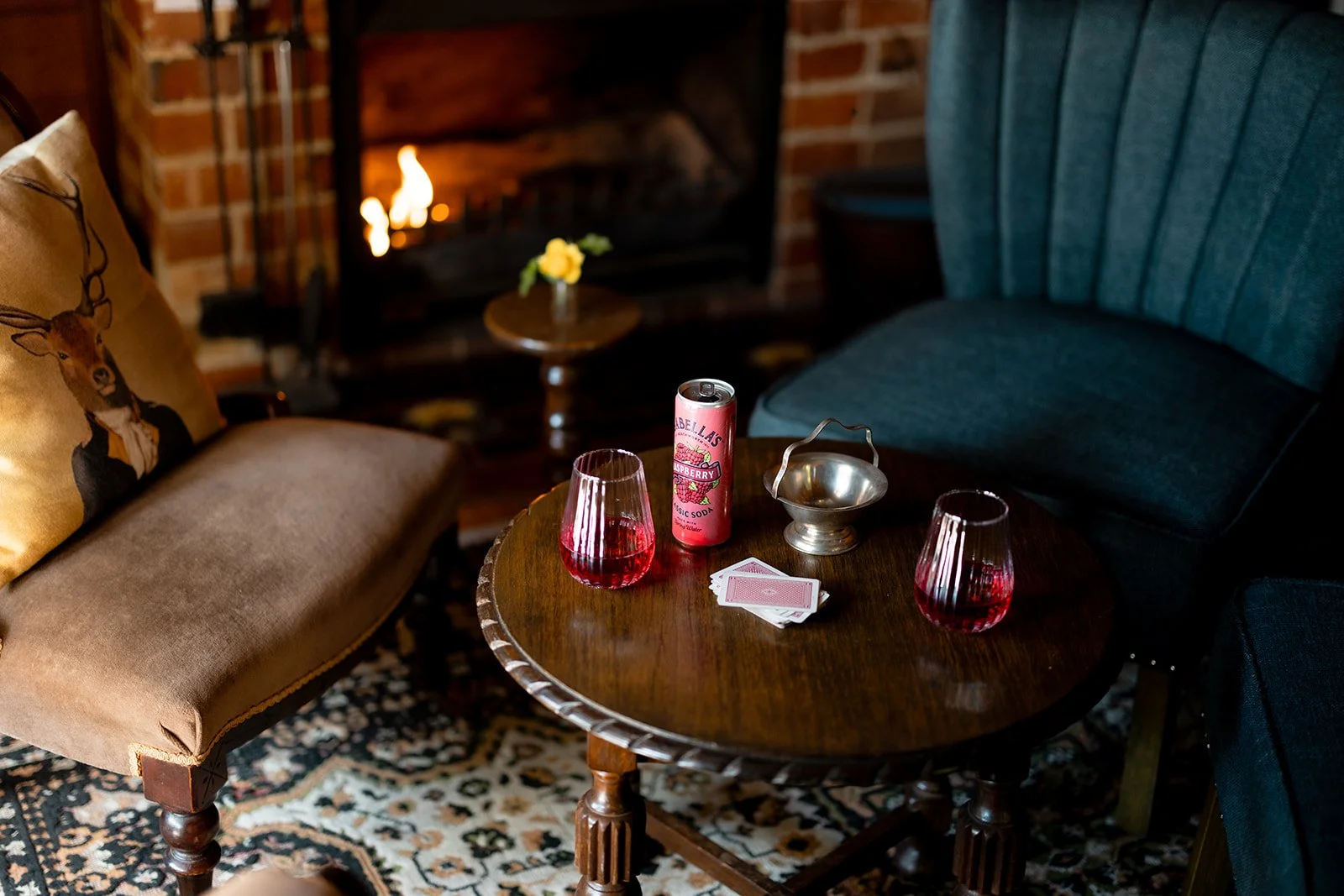 A cozy living room scene with a round wooden table in the center, holding two glasses of pink beverage, a pink soda can, a deck of playing cards, a metal bowl, and a small yellow flower in a vase. To the left is a beige sofa with a deer print pillow,