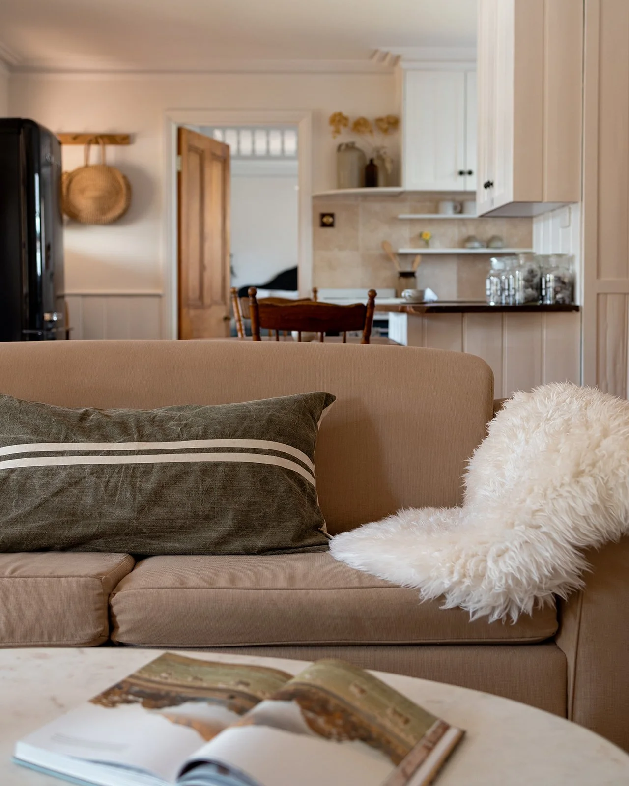 Pickett's Cottage's living room with a beige sofa, dark pillow, white fluffy blanket, open magazine, and view of kitchen and dining area in background.