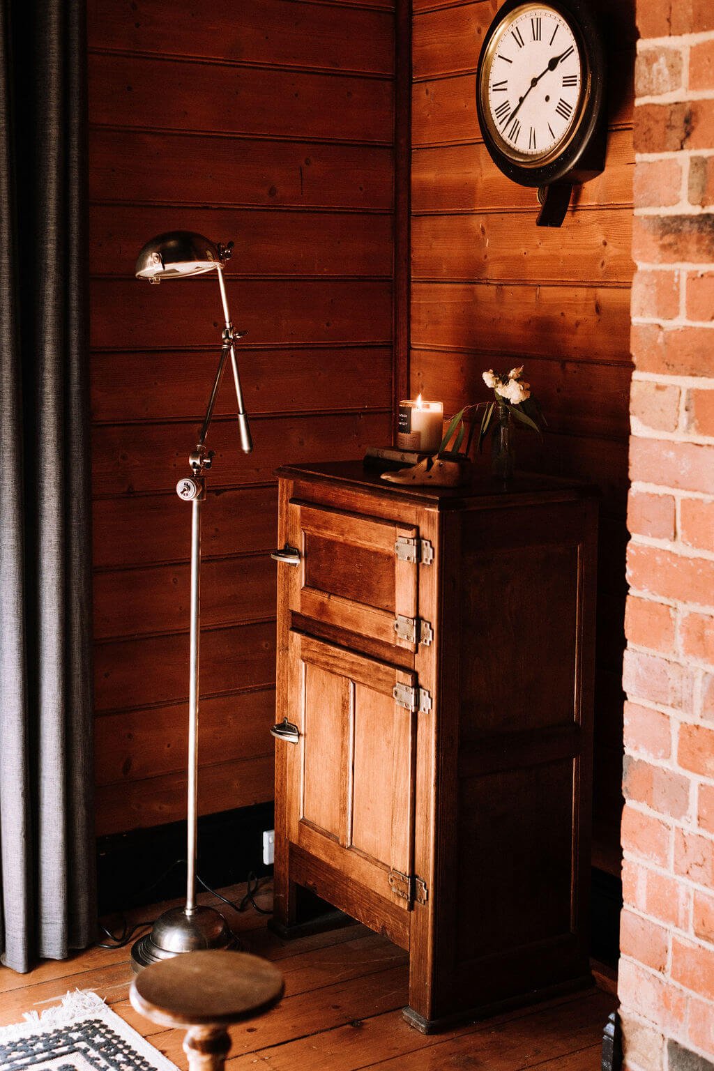 The Inn Beechworth, Luxury Accommodation - Kelly House. Cozy rustic room with wooden walls and floor. A vintage wooden cabinet stands next to a brick wall. A metal floor lamp is beside it. A clock with Roman numerals hangs on the wall above.