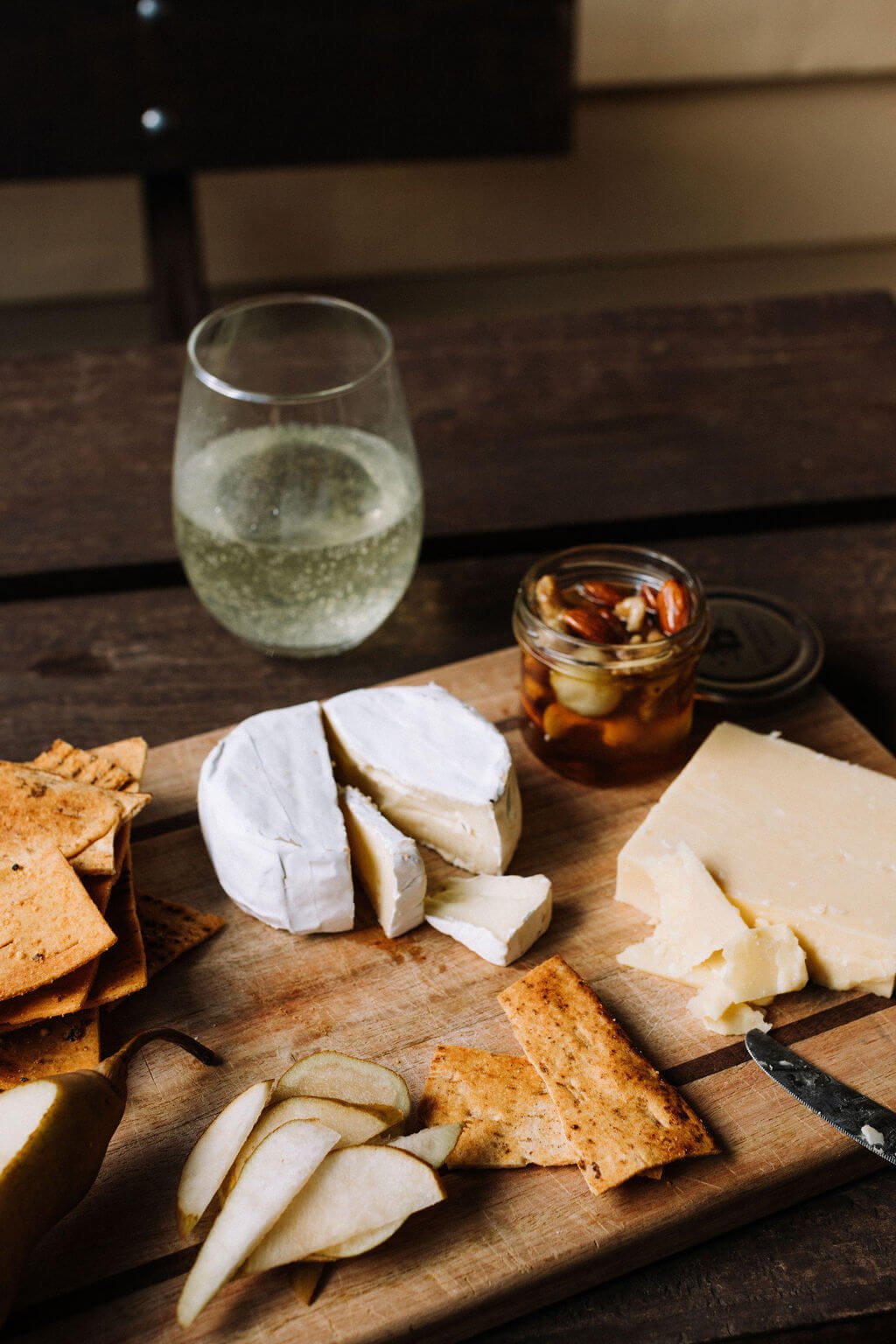 The Inn Beechworth, Luxury Accommodation - Kelly House. Charcuterie board with cheeses, crackers, pear slices, a jar of honey with nuts, and a glass of white wine on a wooden surface.
