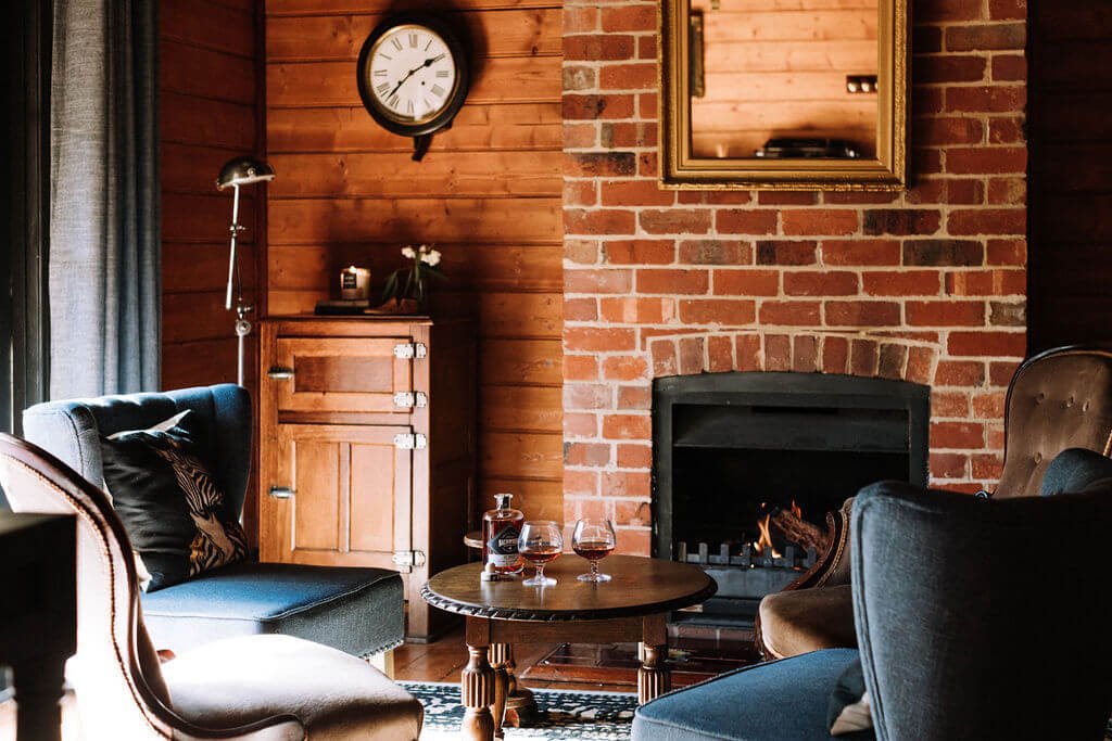 The Inn Beechworth, Luxury Accommodation - Kelly House. Whiskey Room with brick fireplace, wood walls, round table with glassware, vintage clock, and cushioned chairs.