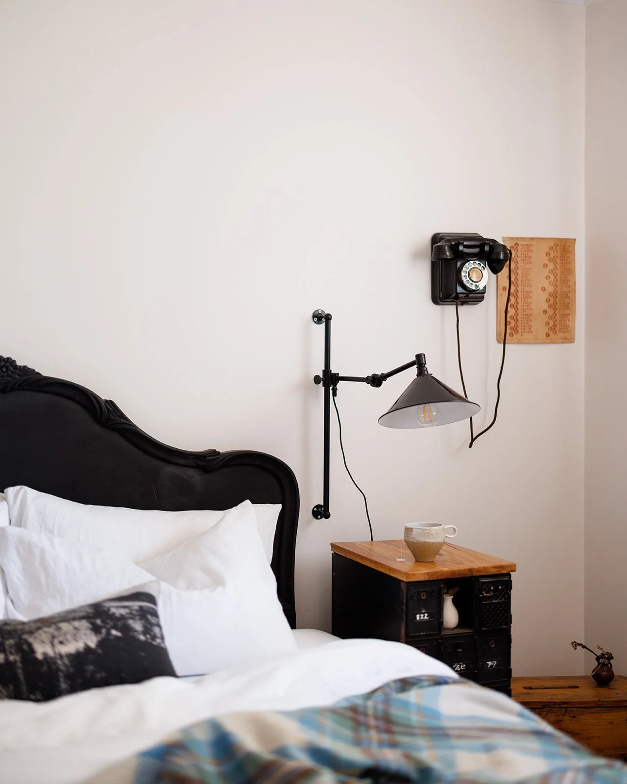 A bedroom with a white wall, black ornate bed frame, white pillows, a black and beige pillow, and a colorful striped duvet. Next to the bed is a black nightstand with a wooden top, a beige cup, and a white vase. Above the nightstand, a vintage black 