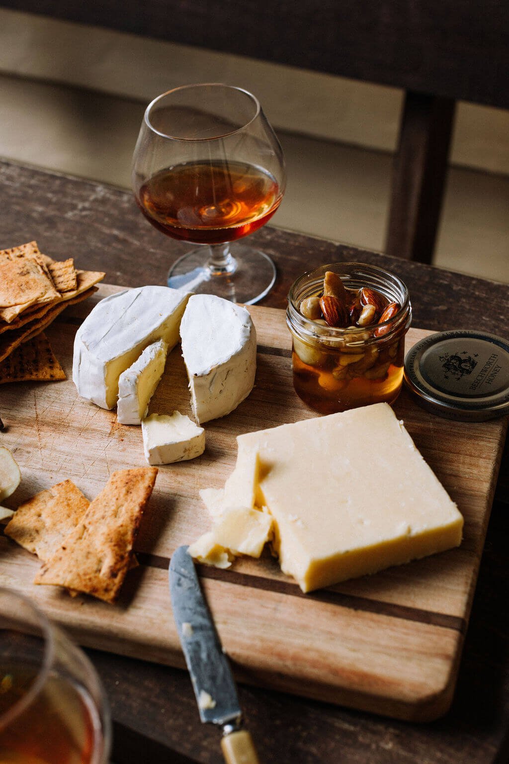 The Inn Beechworth, Luxury Accommodation - Kelly House. Cheese platter with brie, cheddar, crackers, nuts in honey, and a glass of cognac on a wooden board.