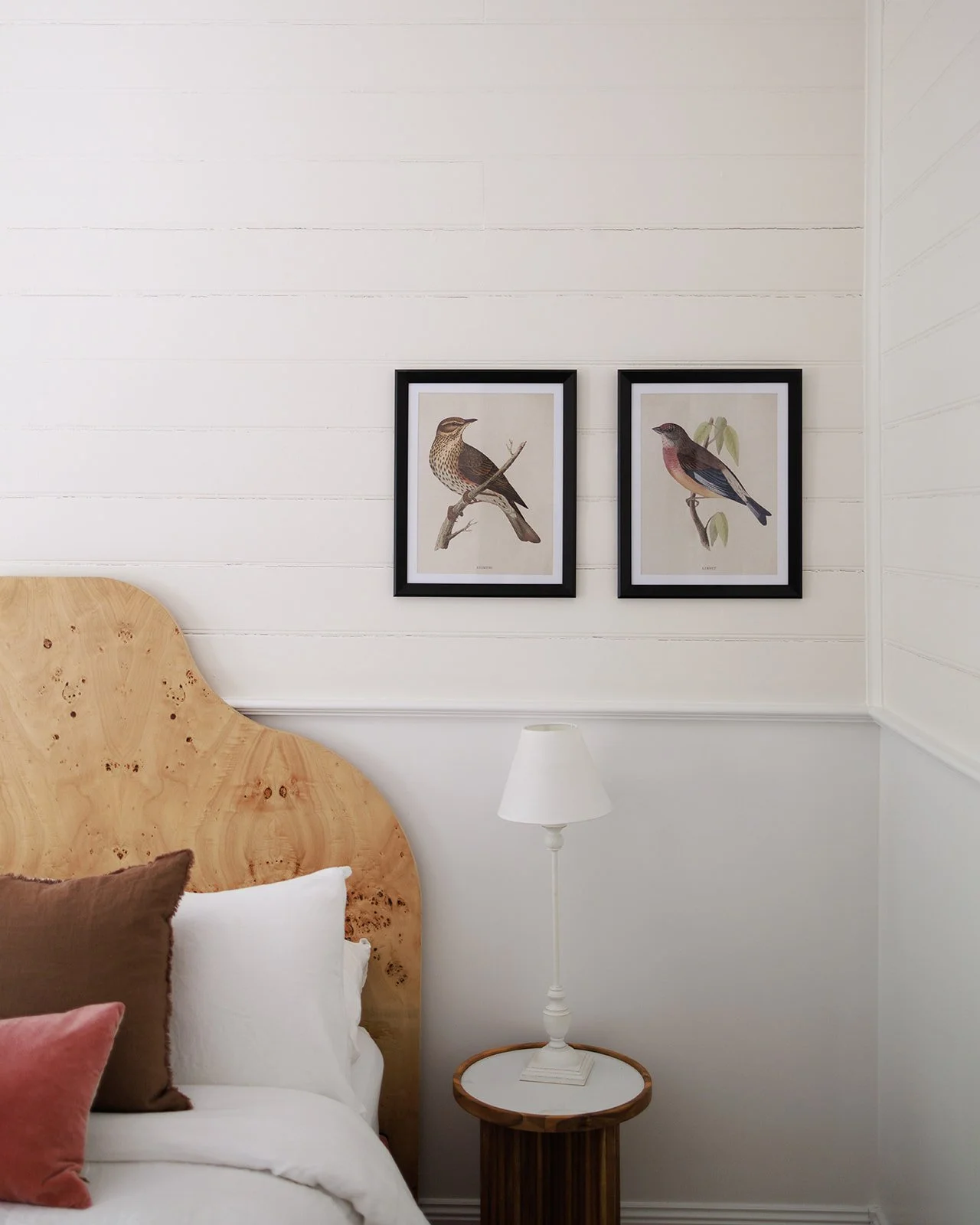 Close-up of a bedroom corner with a wooden headboard, pillows, a white bedside lamp, and two framed bird illustrations on white shiplap wall.