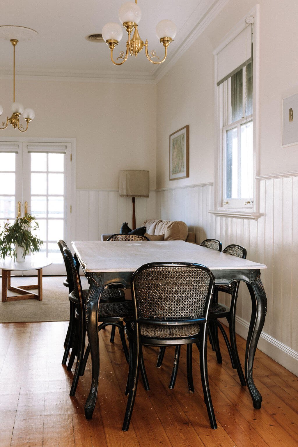 The Inn Beechworth, Luxury Accommodation - Kelly House. Dining room with a white table, black chairs, wooden floor, beige sofa, lamp, round coffee table with plant, wall art, and pendant lights.