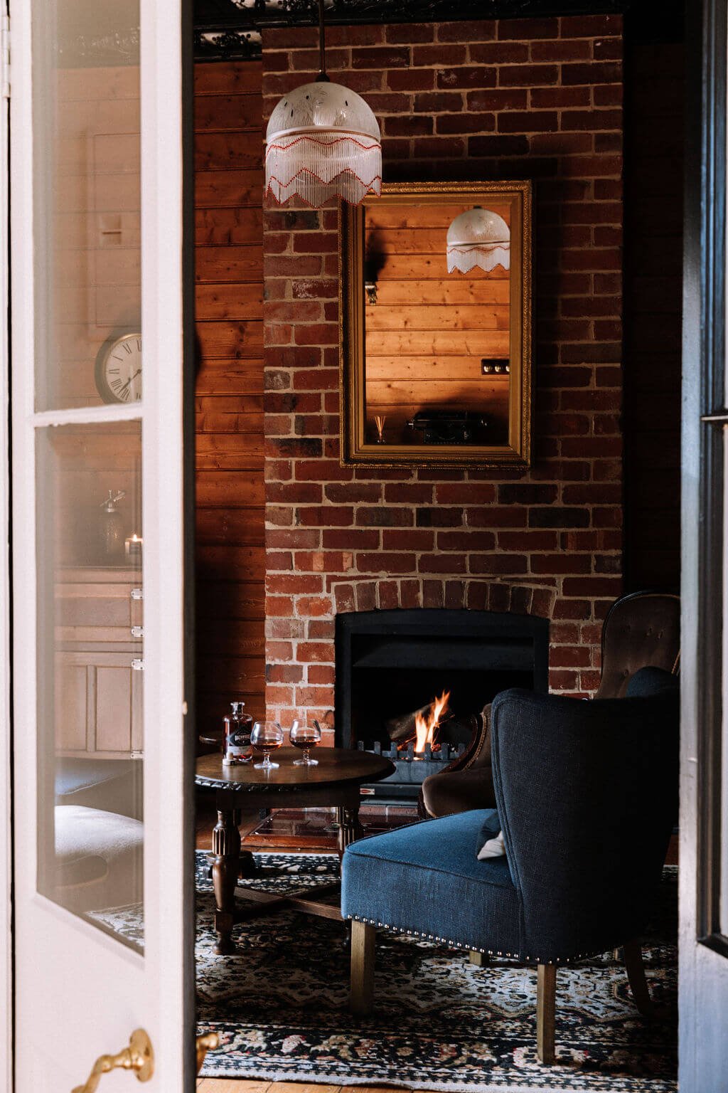 The Inn Beechworth, Luxury Accommodation - Kelly House. Cozy living room with a brick wall, lit fireplace, dark blue armchair, and wooden coffee table. A mirror hangs above the fireplace, reflecting a pendant lamp and wood-paneled walls.