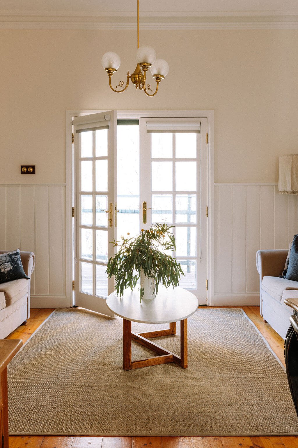 The Inn Beechworth, Luxury Accommodation - Kelly House. Cozy living room with French doors, a chandelier, a round coffee table with a plant, beige sofas, and wooden flooring.