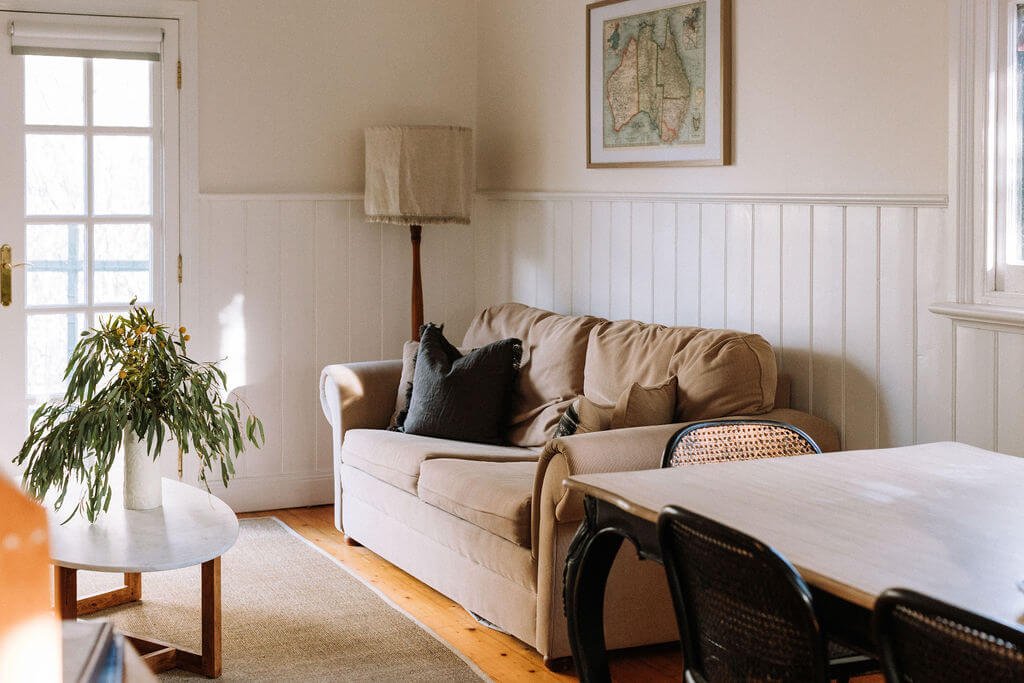 The Inn Beechworth, Luxury Accommodation - Kelly House. Cozy living room with beige couch, decorative pillows, wooden floor, white panelling, framed map, tall lamp, plant on the coffee table, and dining table with chairs.