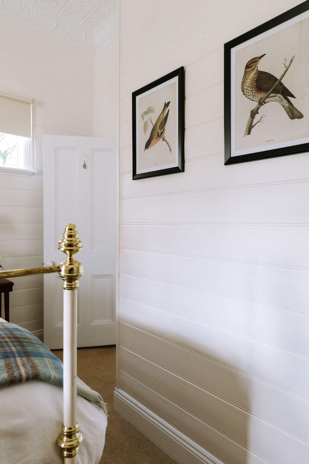 The Inn Beechworth, Luxury Accommodation - Kelly House. Bedroom interior with framed bird art on white paneled wall, brass bedpost, and a white door.
