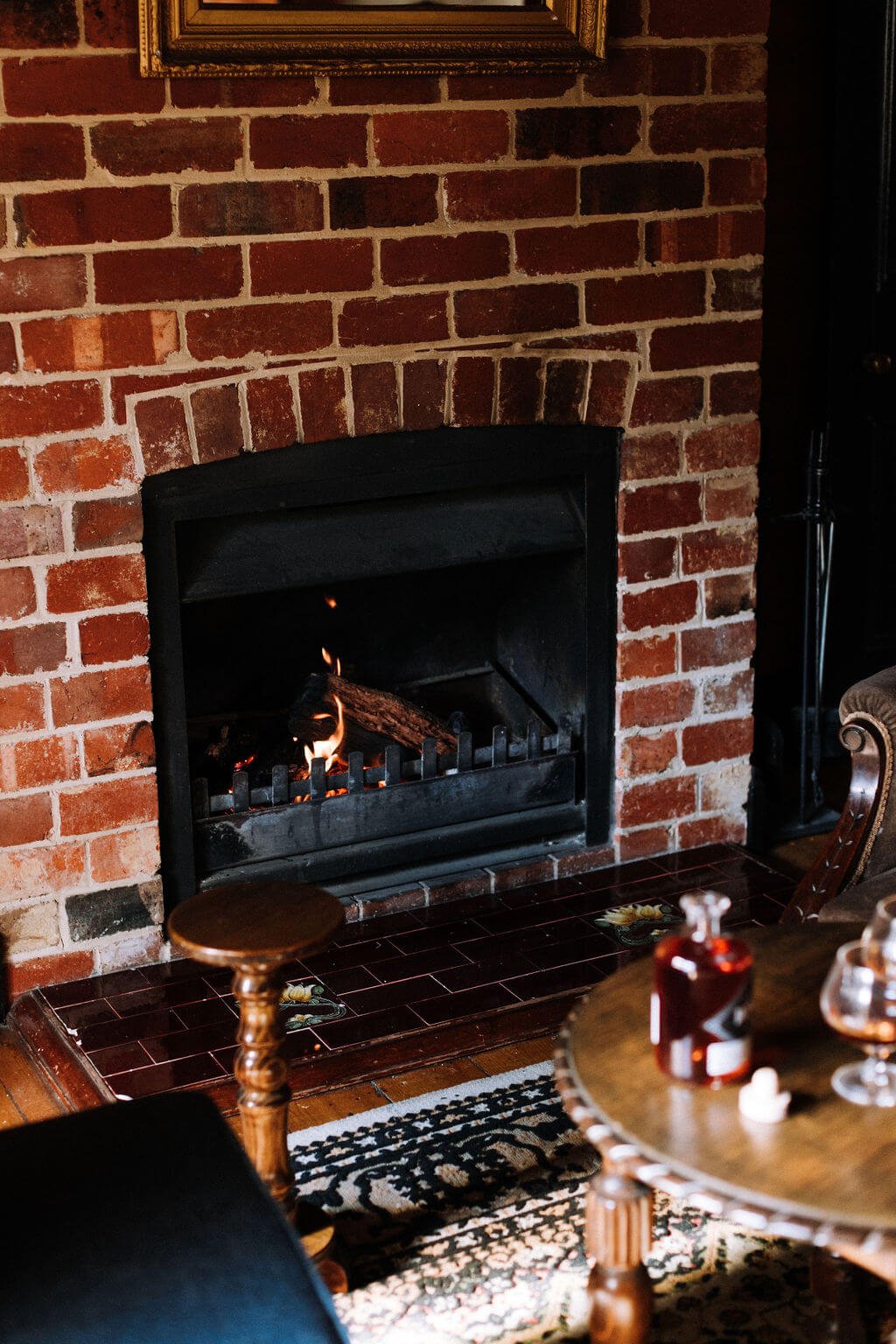 The Inn Beechworth, Luxury Accommodation - Kelly House. Cozy living room with a lit fireplace set in a red brick wall, surrounded by wooden furniture and a patterned rug, with drinks on a table nearby.