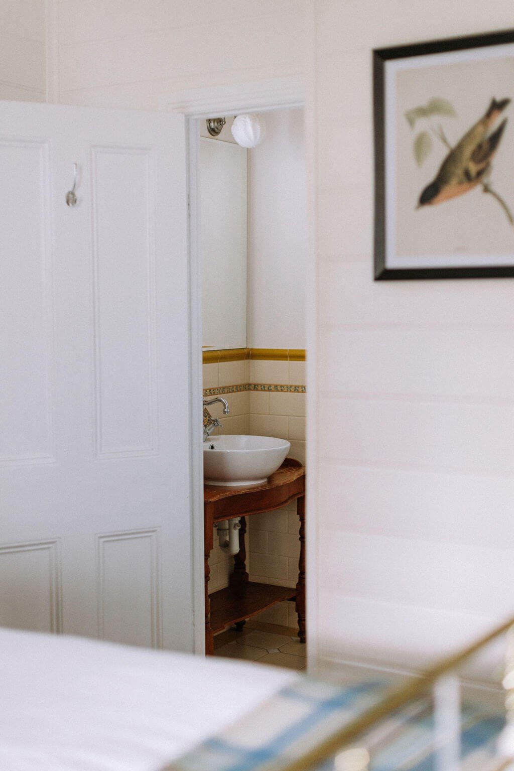 The Inn Beechworth, Luxury Accommodation - Kelly House. View of a partially open white door revealing a bathroom sink on a wooden table with beige tiled walls. A framed bird illustration hangs on an adjacent wall.