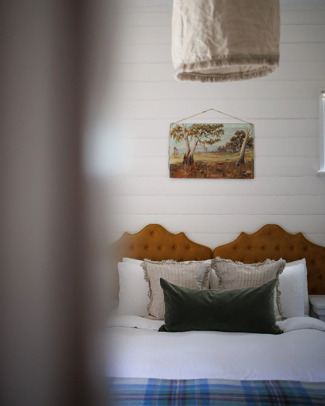 A cozy bedroom with a white wall, a framed landscape painting, a wooden headboard, and pillows, with a beige hanging fabric partially visible in the foreground.