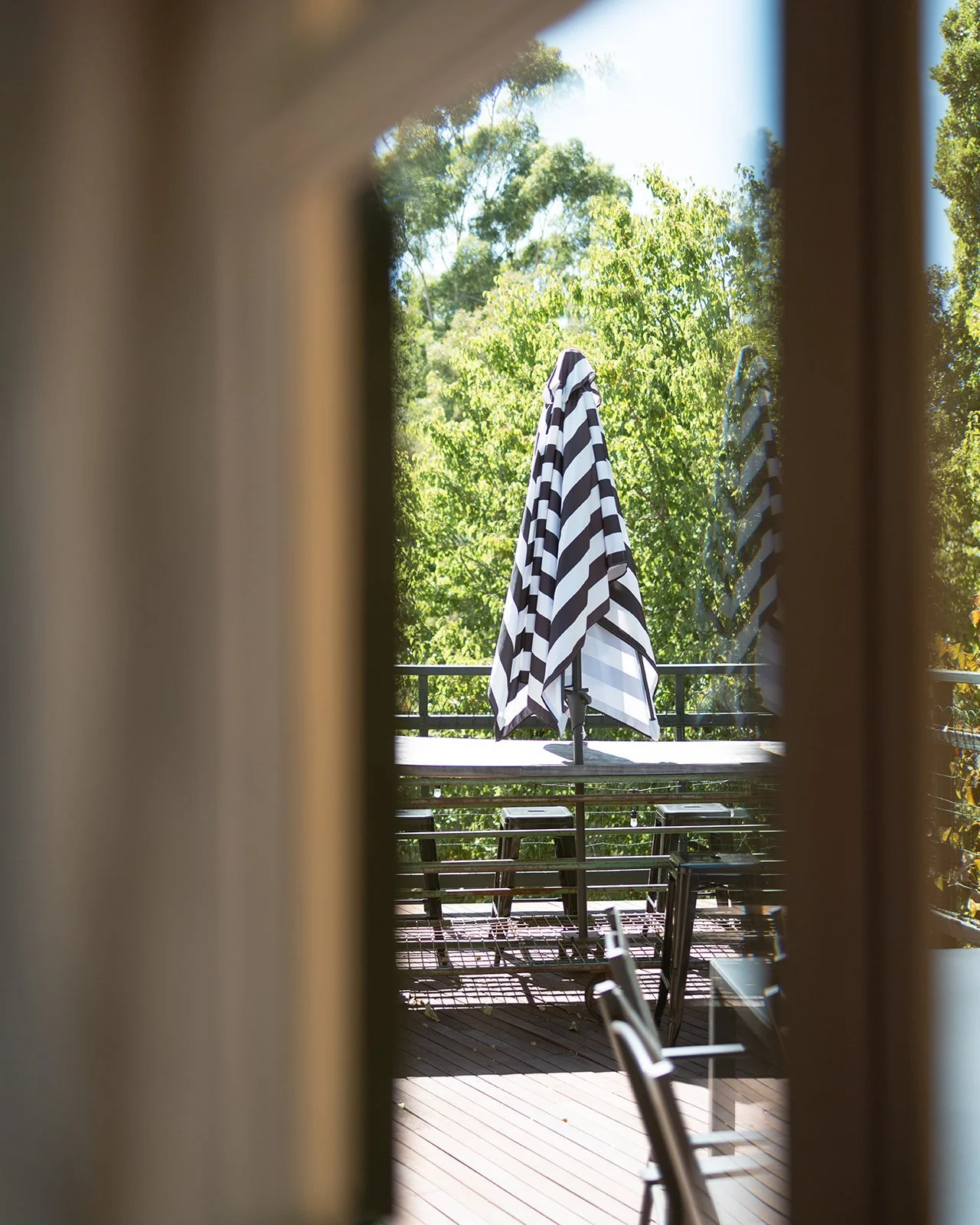 View through a window showing an outdoor patio with black and white striped umbrellas, chairs, and a railing, surrounded by green trees and clear sky.