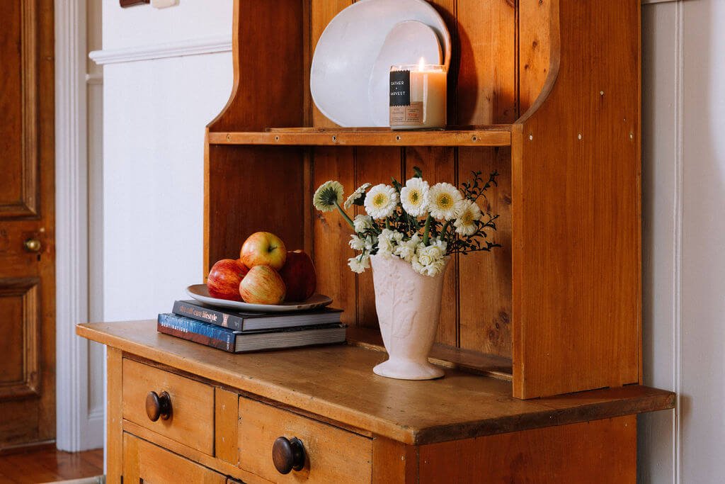 The Inn Beechworth, Luxury Accommodation - Kelly House. Wooden hutch with a vase of white flowers, a plate of apples on books, and a lit candle.