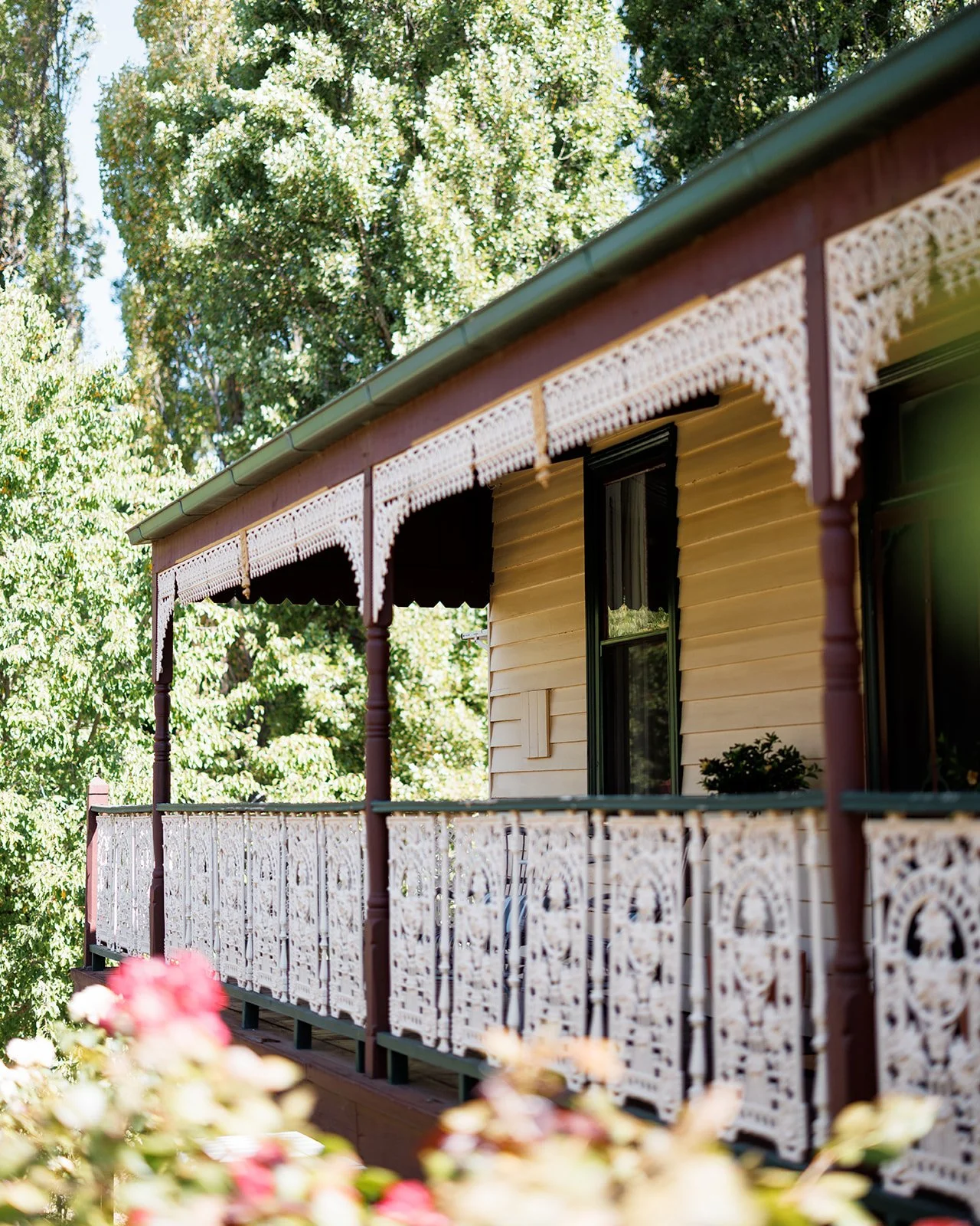 Front porch of a house with an ornate white lace trim on the roof and railing, yellow siding, and a green roof, surrounded by leafy green trees.