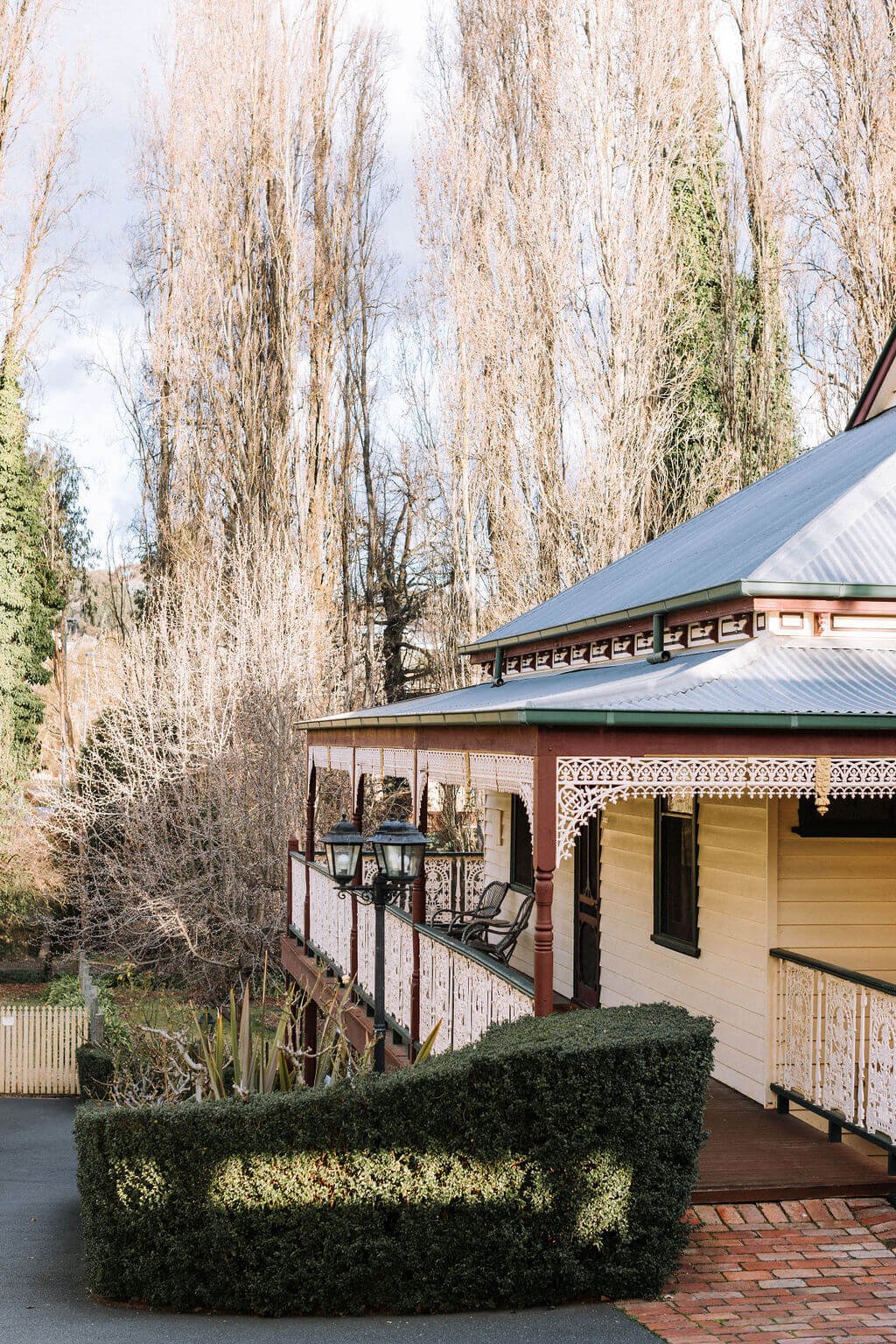 The Inn Beechworth, Luxury Accommodation - Kelly House. Victorian-style house with ornate porch railing, surrounded by bare trees and shrubs, set in a countryside setting.