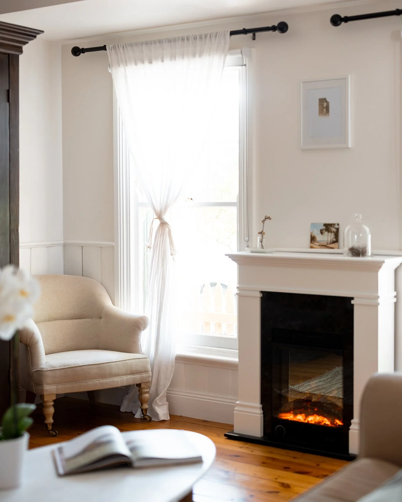Pickett's Cottage has a bright living room with white walls, a beige armchair, a white fireplace with a digital fire display, a window with sheer curtains tied back, and minimal decorative items on the mantel and wall.