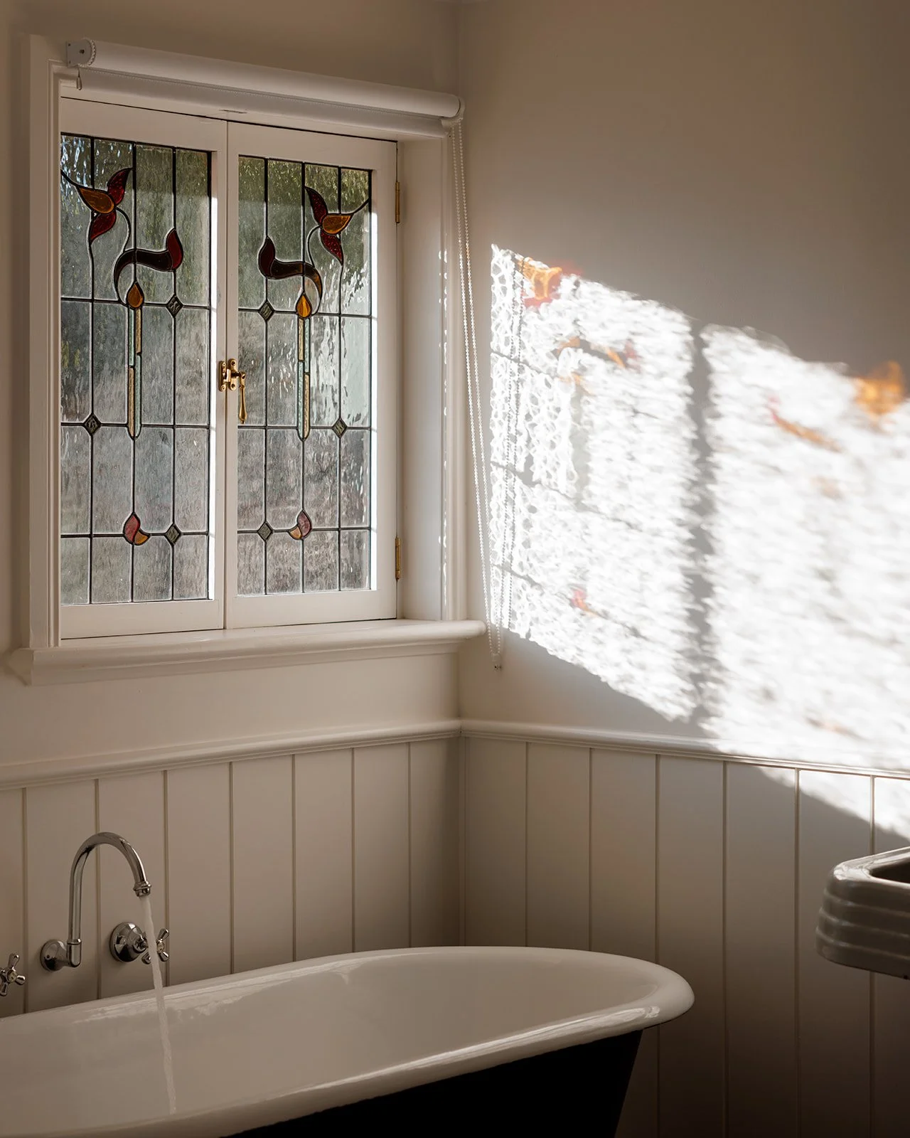A bathroom with a stained glass window, a bathtub with running water, and sunlight casting patterned shadows on the wall.