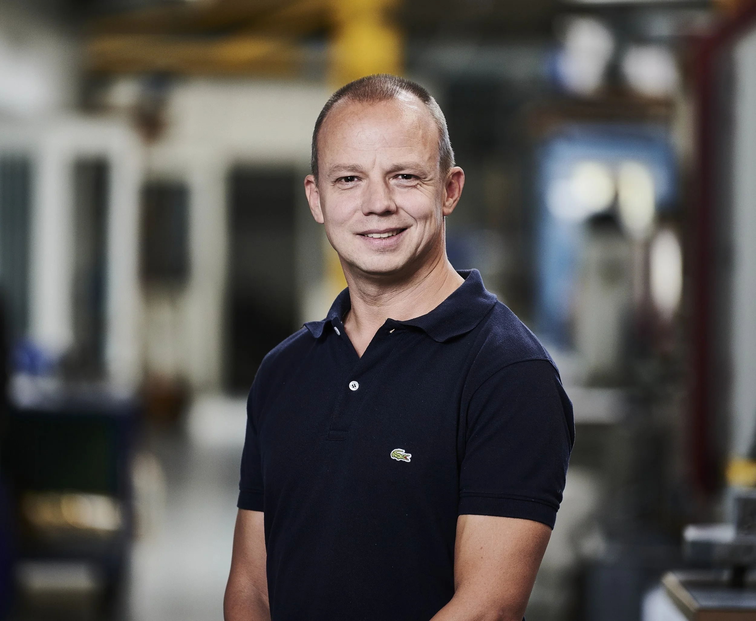 Portræt af en mand med kort hår, iført en navyblå polo i en industriel eller værksteds-lignende omgivelser.