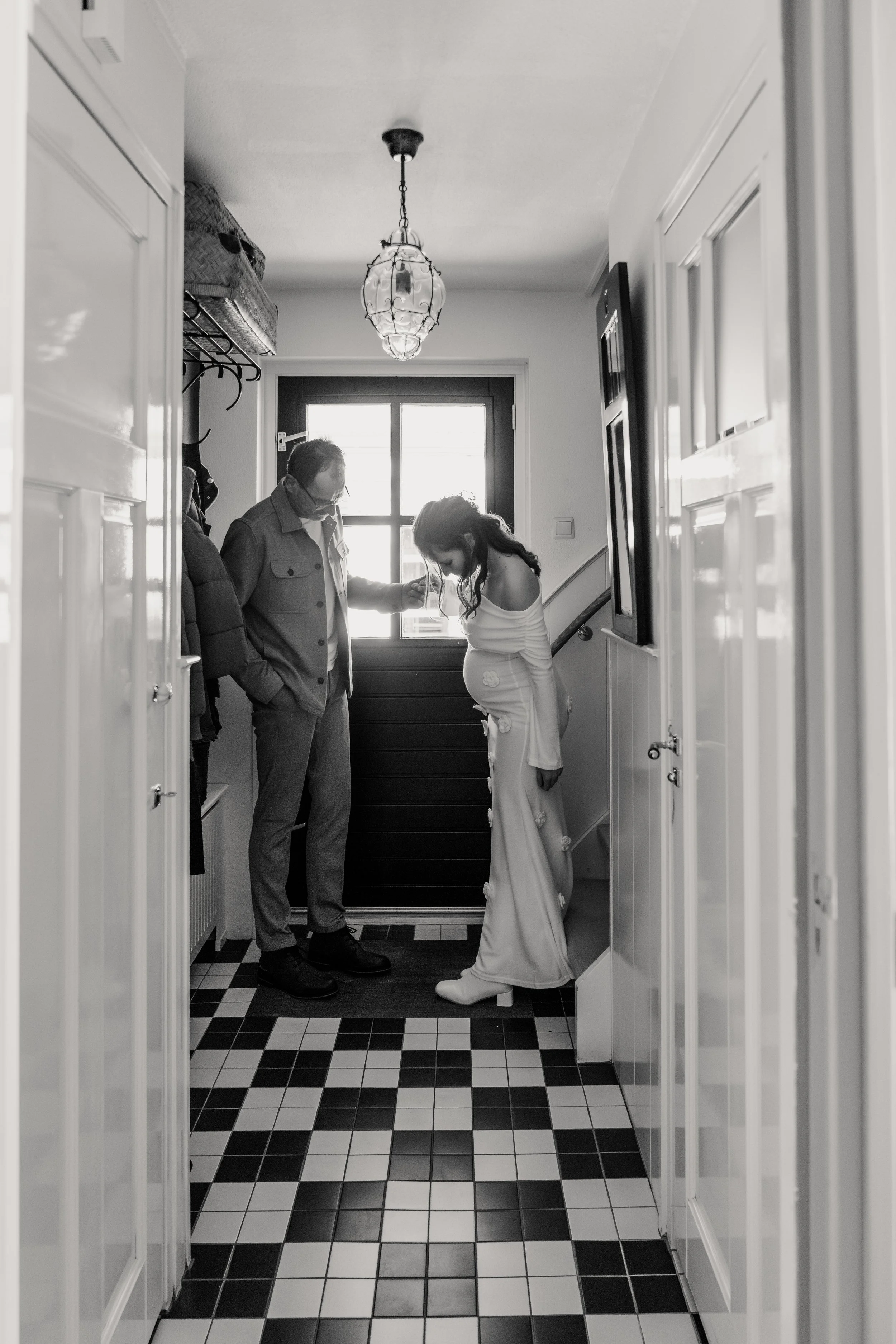 Een zwangere vrouw in een witte jurk bukt zich en aait haar buik terwijl twee mannen, een draagt een masker en de ander heeft een bril, haar liefdevol aankijken in een hal met zwart-wit tegelvloer.